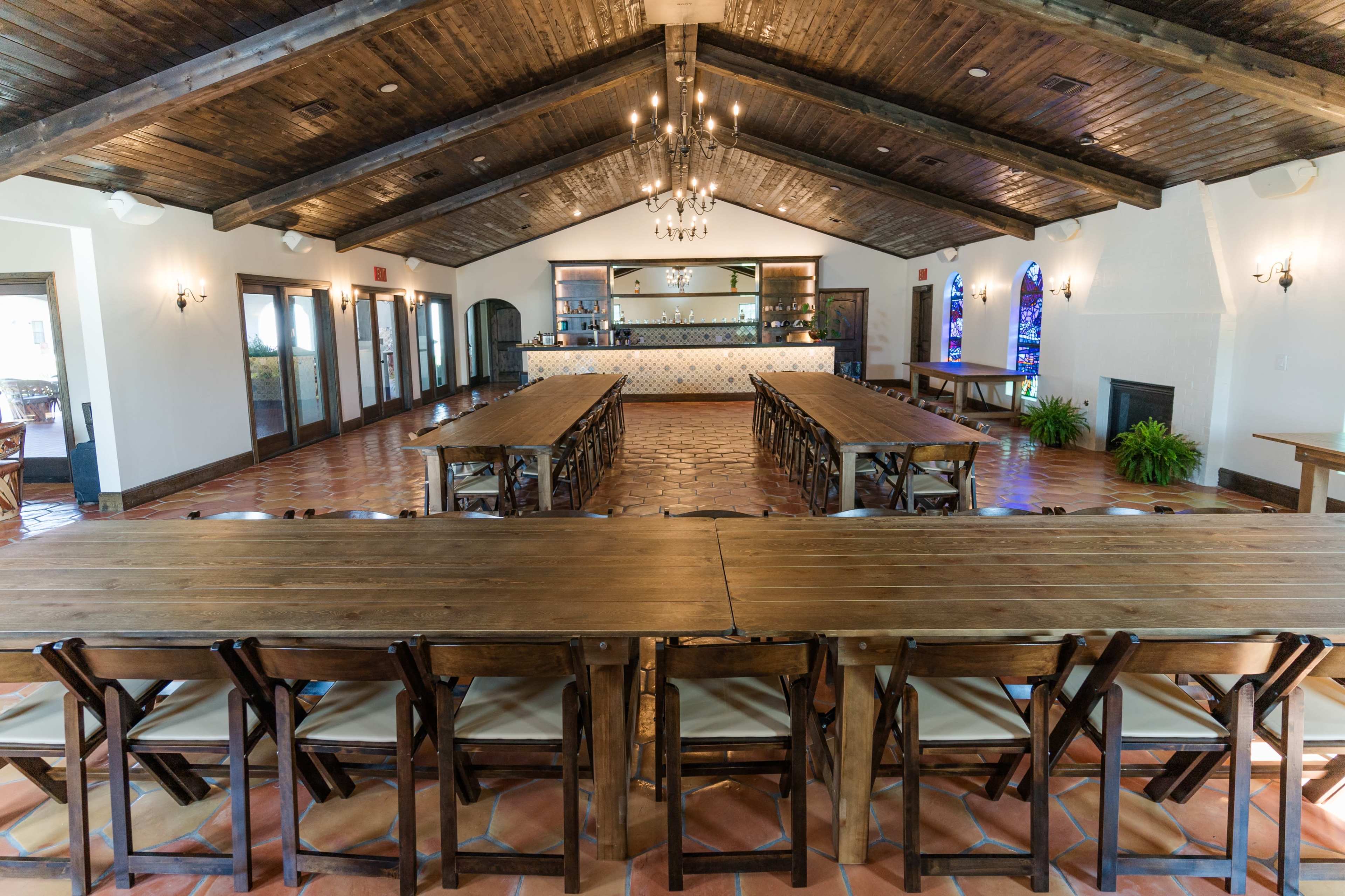 A spacious dining area with wooden tables arranged in a long configuration, featuring a bar with shelves at the back and pendant lighting above.