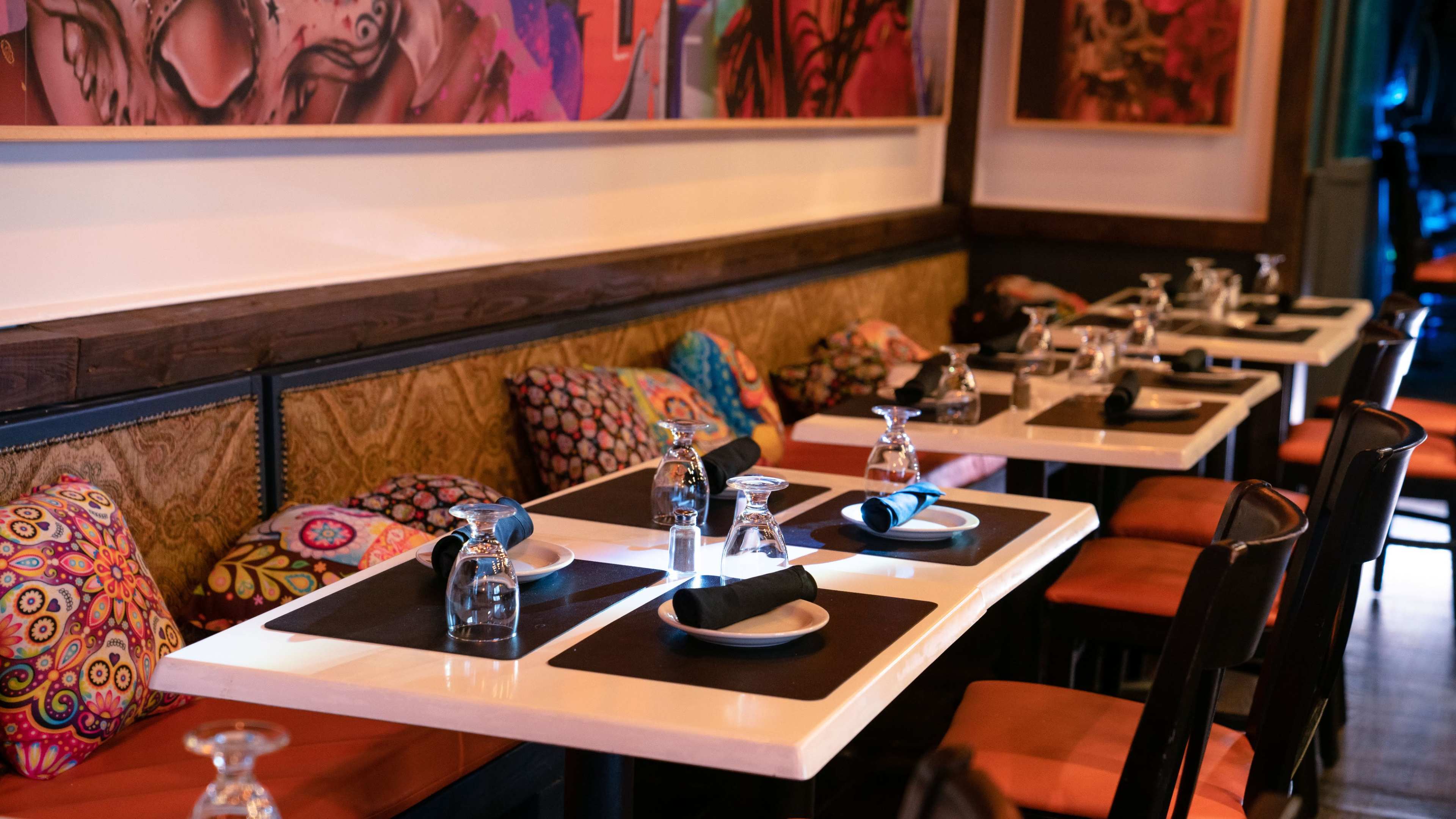 The image shows a restaurant interior with tables set for dining, featuring colorful cushions and water carafes on each table.