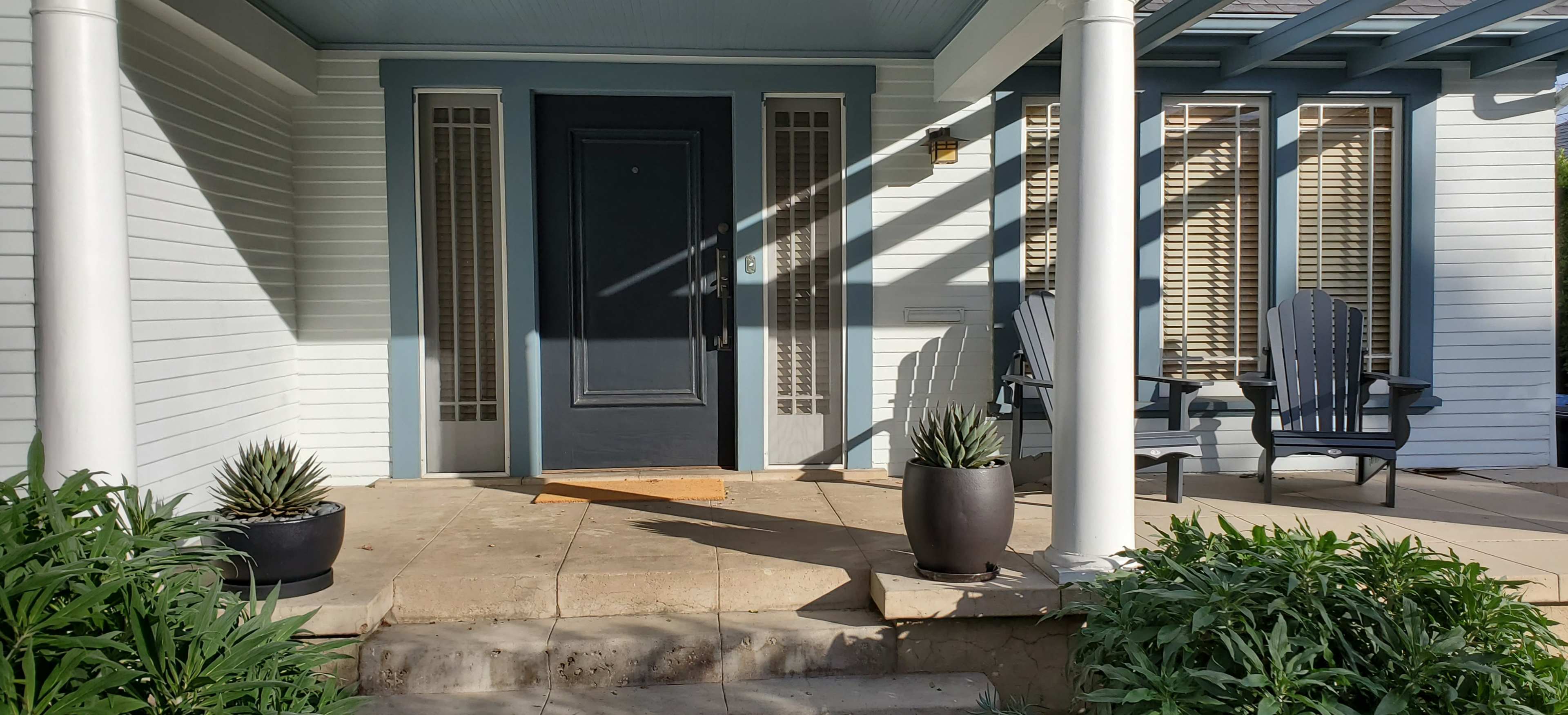 The entrance features a dark door surrounded by large windows, two potted plants, and two chairs on a porch with contrasting sunlight and shadows.