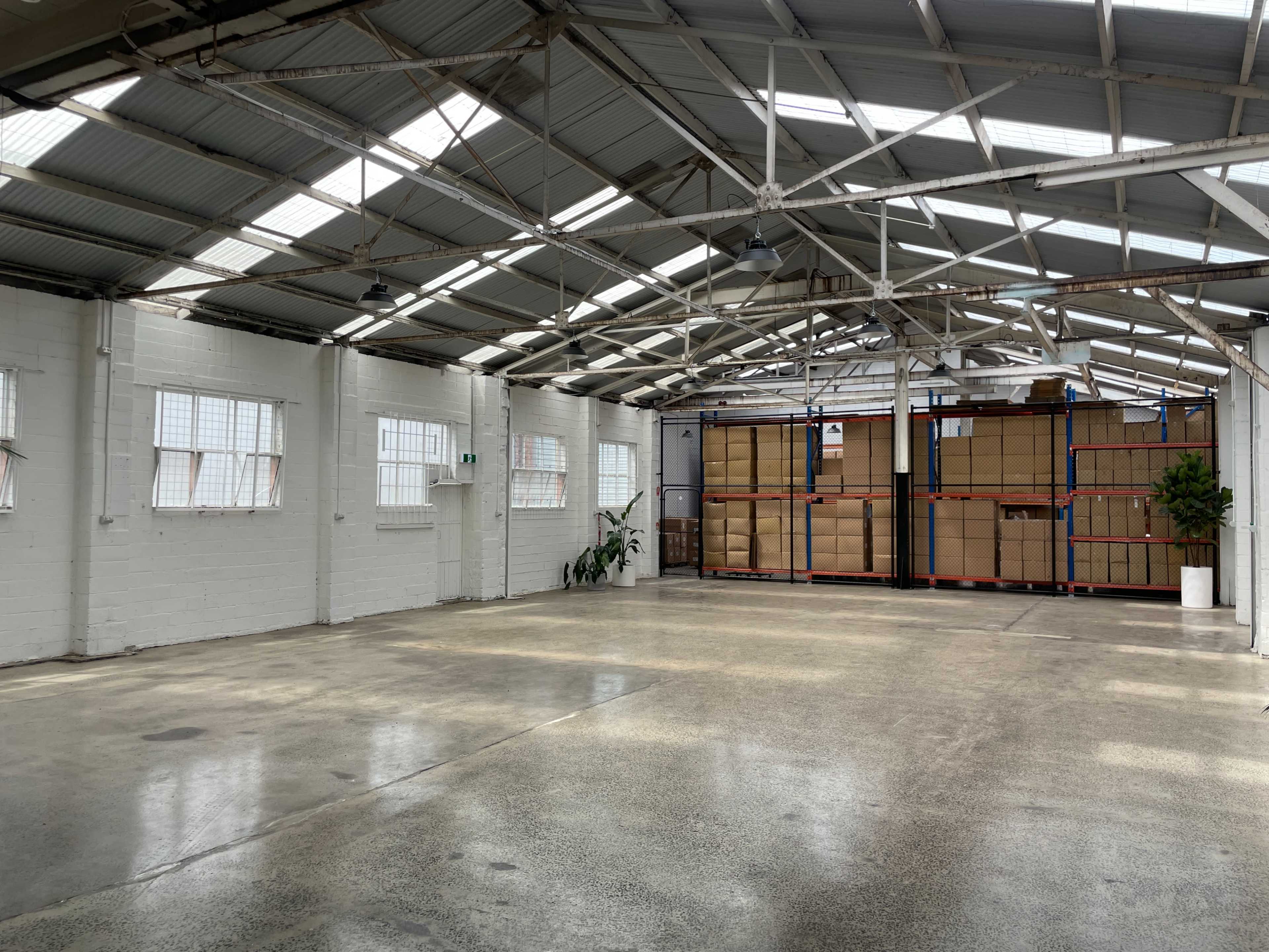 The image shows an empty warehouse with high ceilings, large windows, and shelves filled with cardboard boxes in the back.