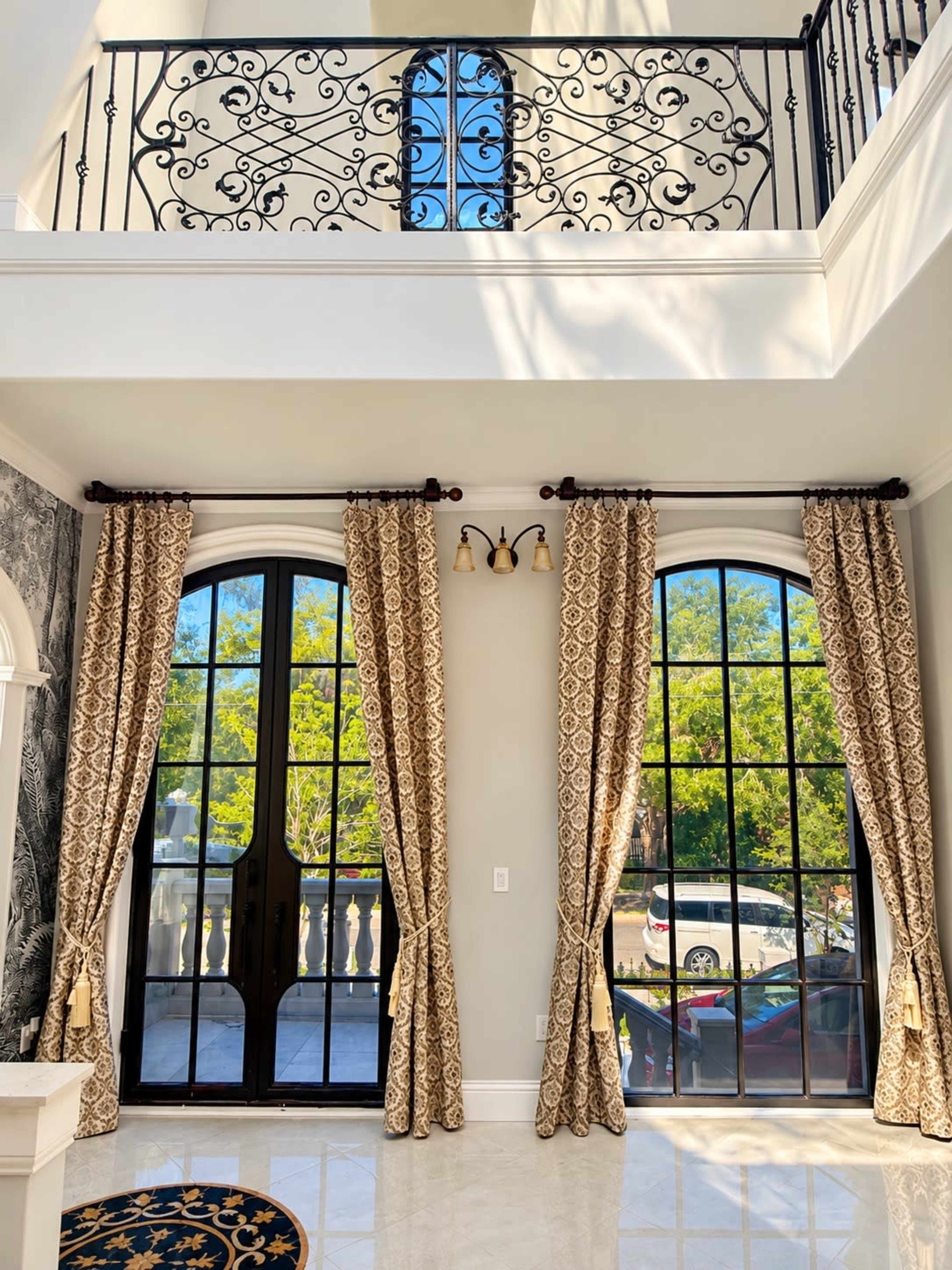 The image shows a spacious interior with large black-framed windows, elegant curtains, and a balcony with ornate iron railing.