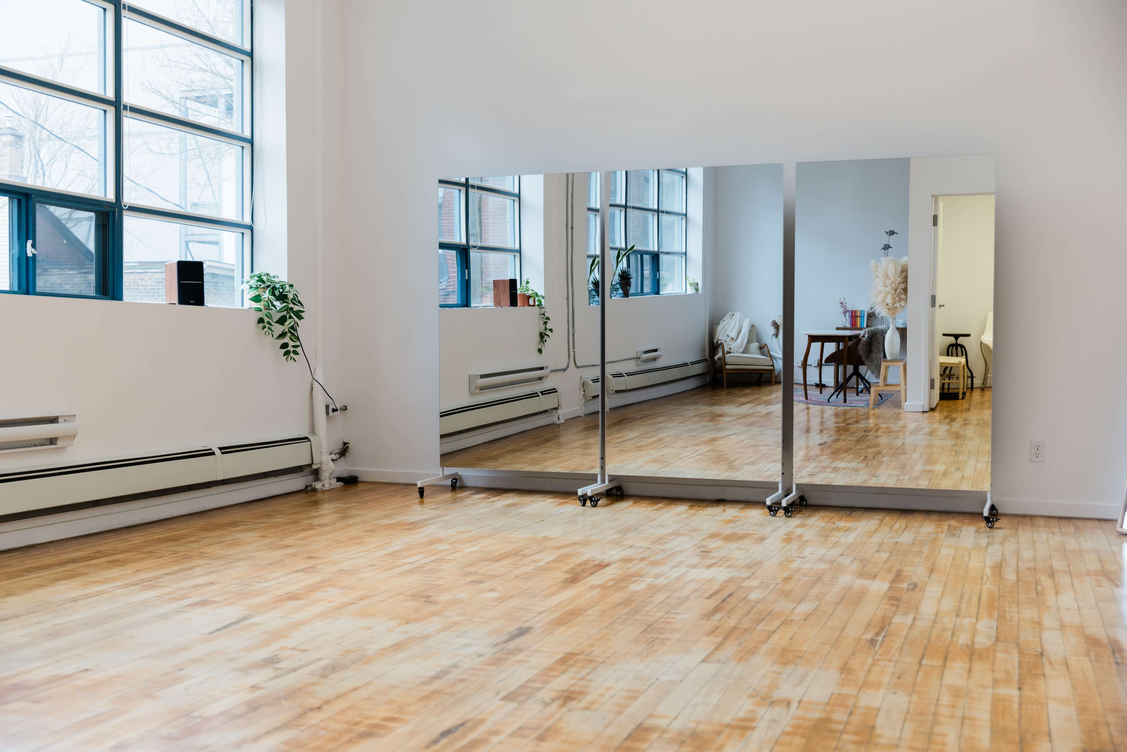 The image shows a bright room with large windows, hardwood floors, and three mirrored panels against a white wall.