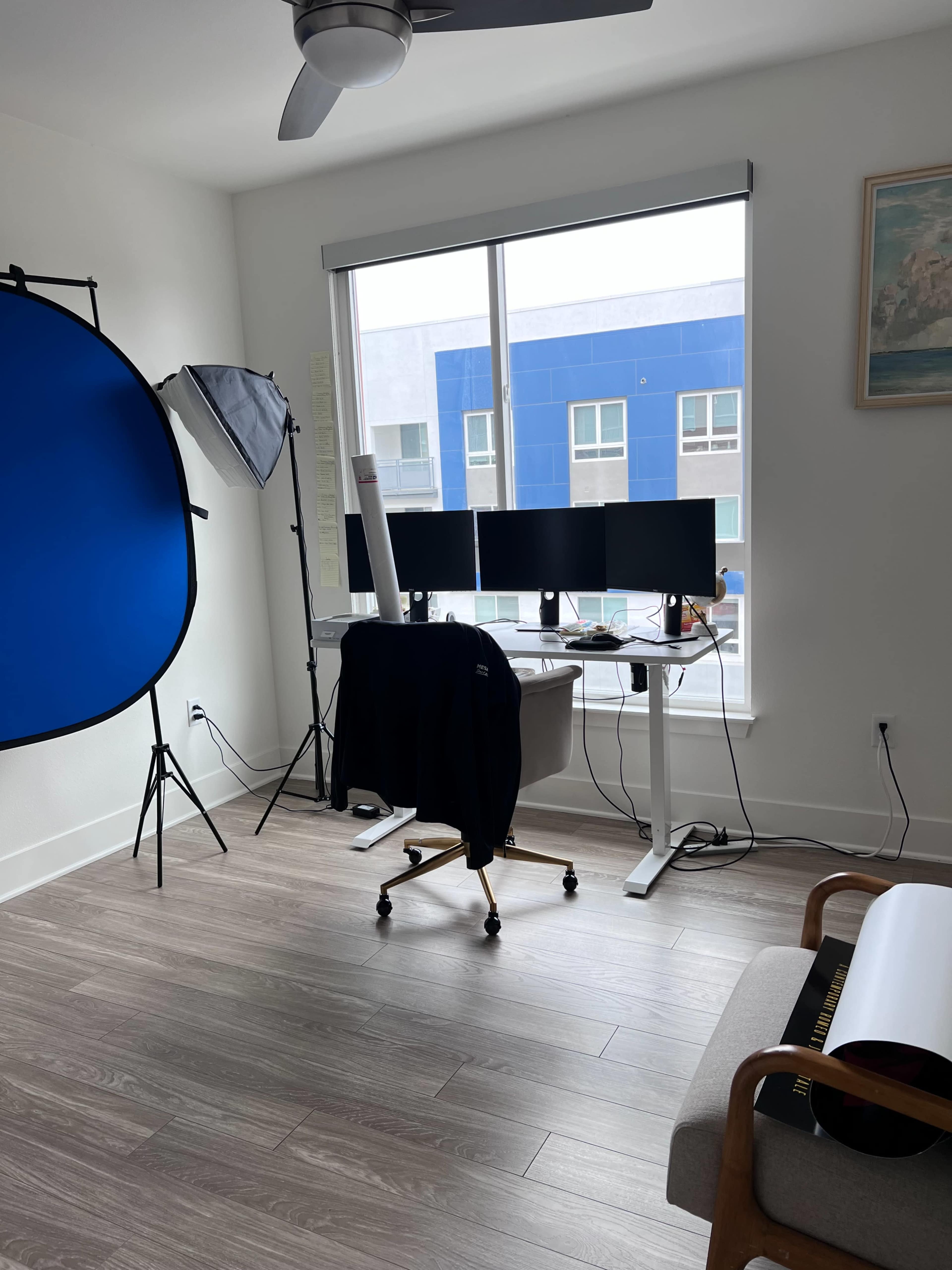 The image shows a well-lit home office with three computer monitors on a height-adjustable desk, a blue backdrop, and a large softbox for lighting.