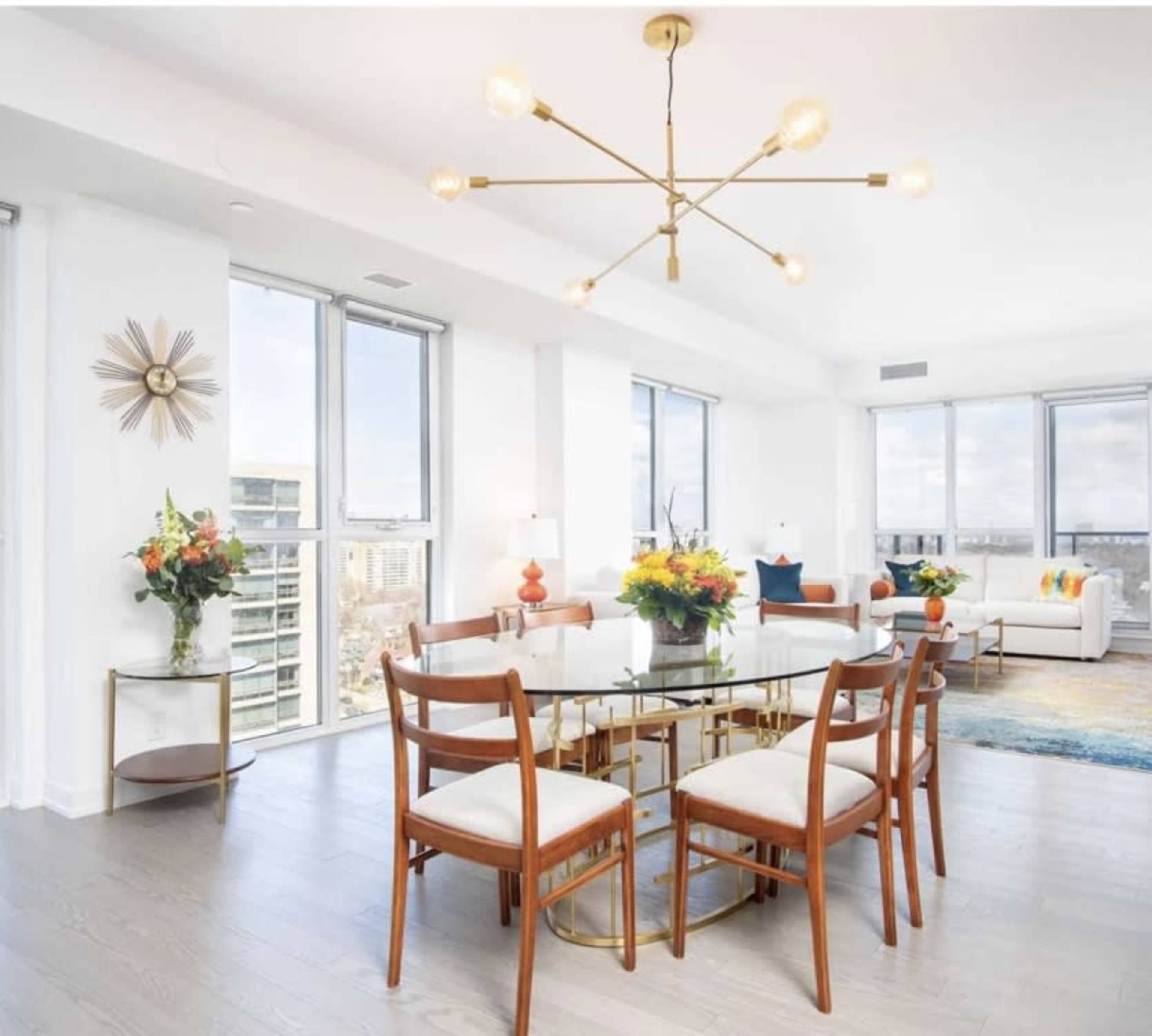 A modern dining area features a circular glass table surrounded by wooden chairs, with large windows offering a view of the city.