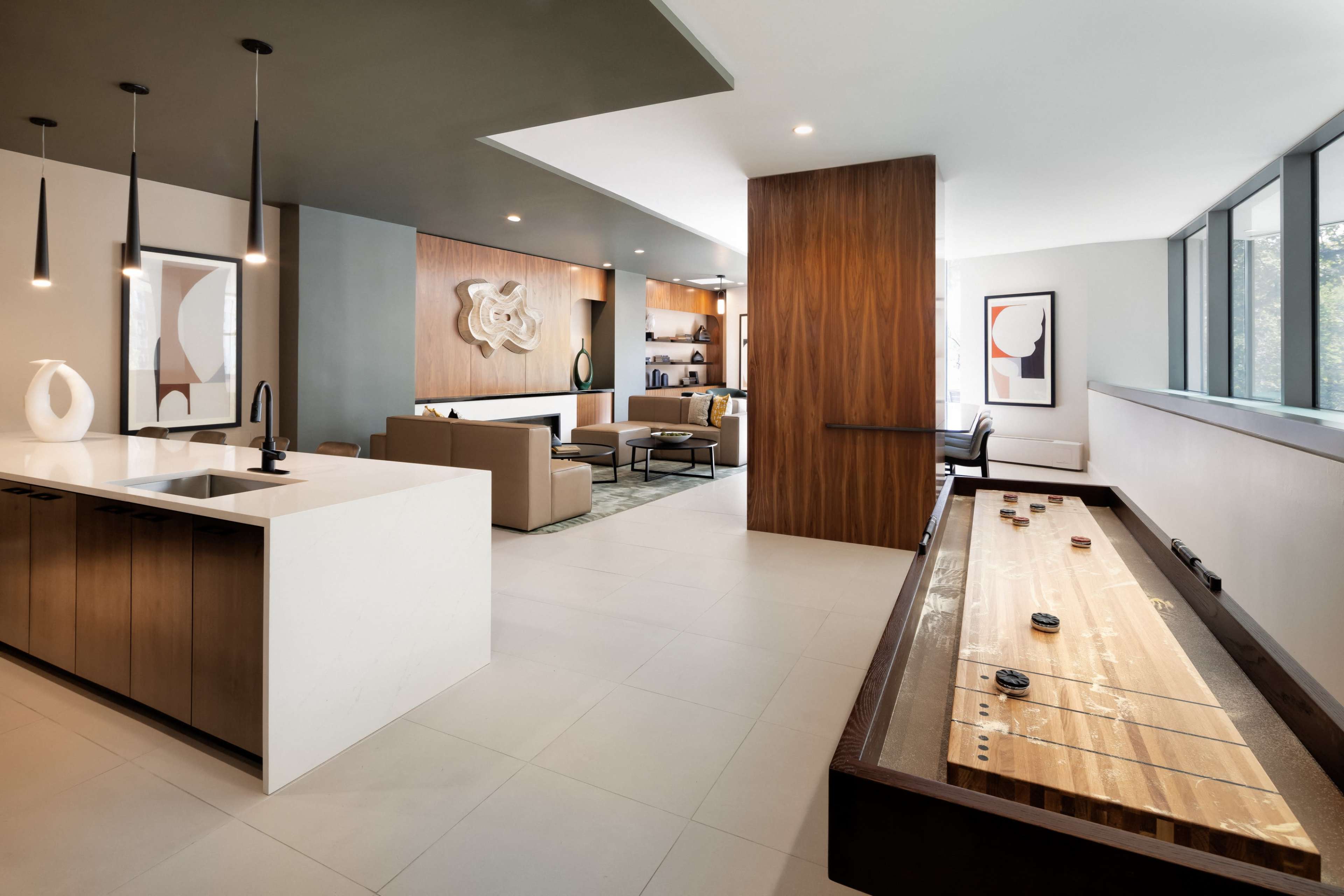 The image shows a modern indoor space featuring a kitchen area with a sink and countertop, a living room with seating, and a shuffleboard table.