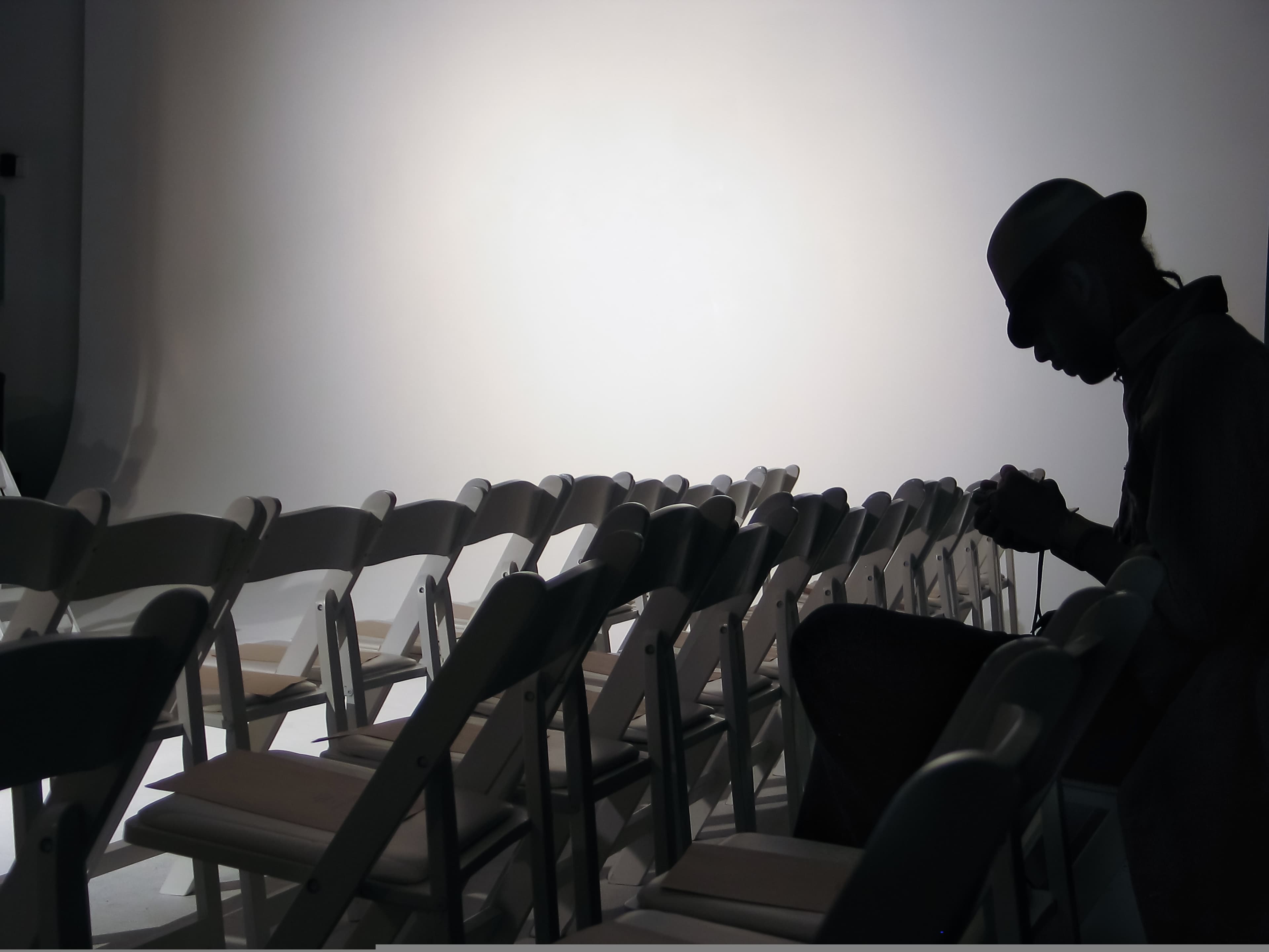 A silhouette of a person wearing a hat sits in front of empty white chairs against a bright background.