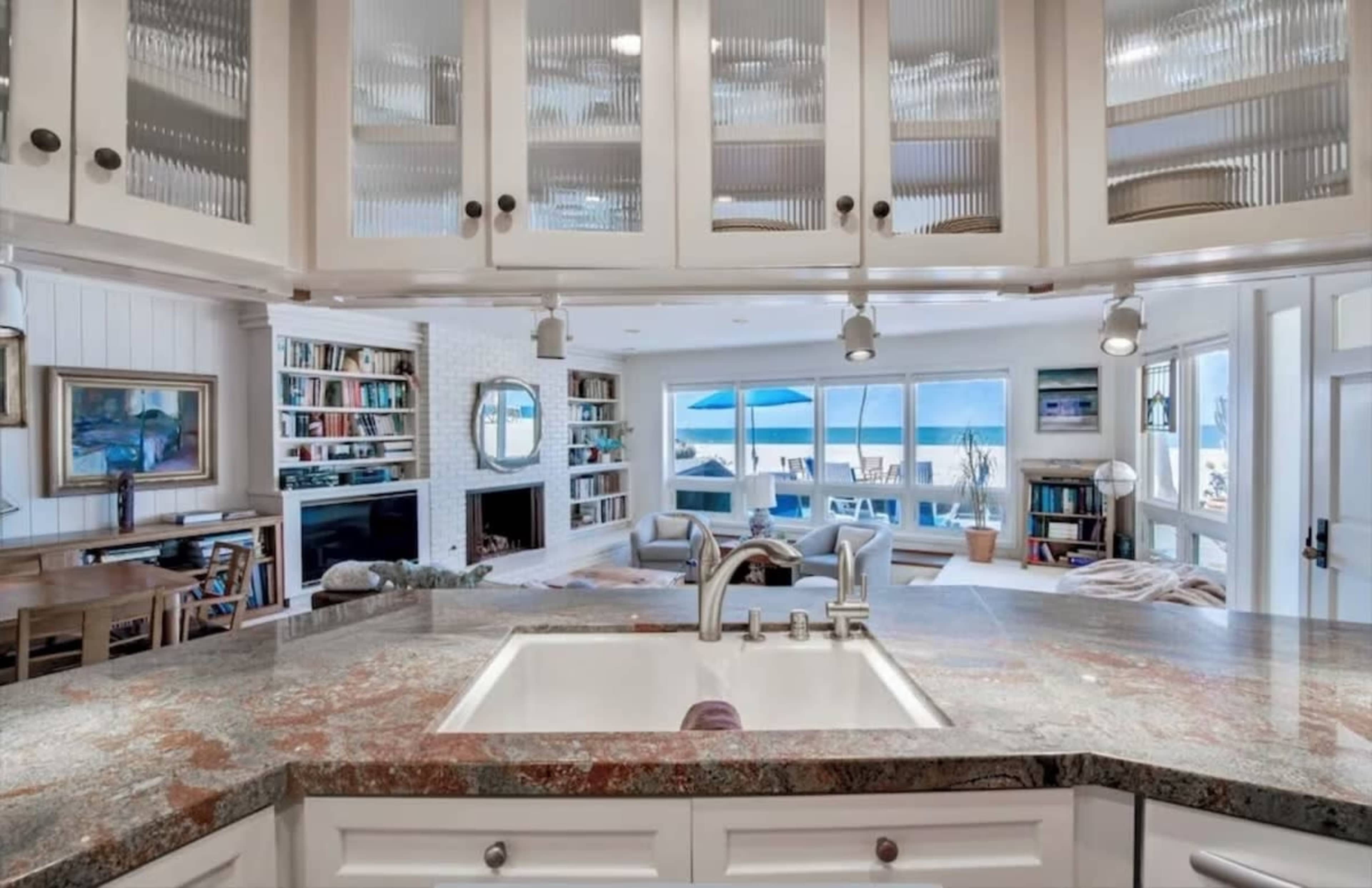 The image shows a modern kitchen with a granite countertop that overlooks a living area with large windows and a view of the ocean.