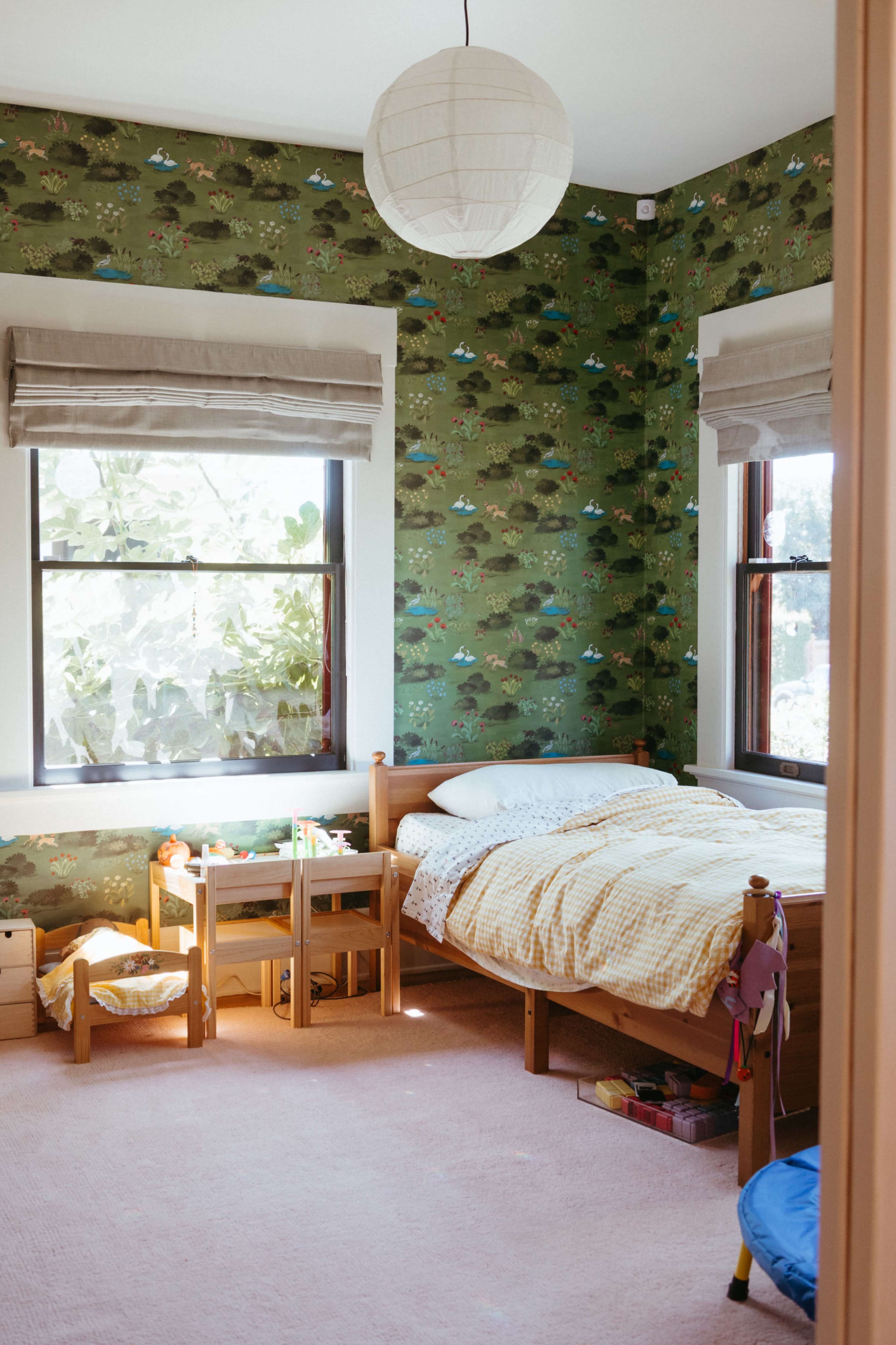 The image shows a children's bedroom with green patterned wallpaper, a bed with yellow bedding, and wooden furniture including small tables and storage.