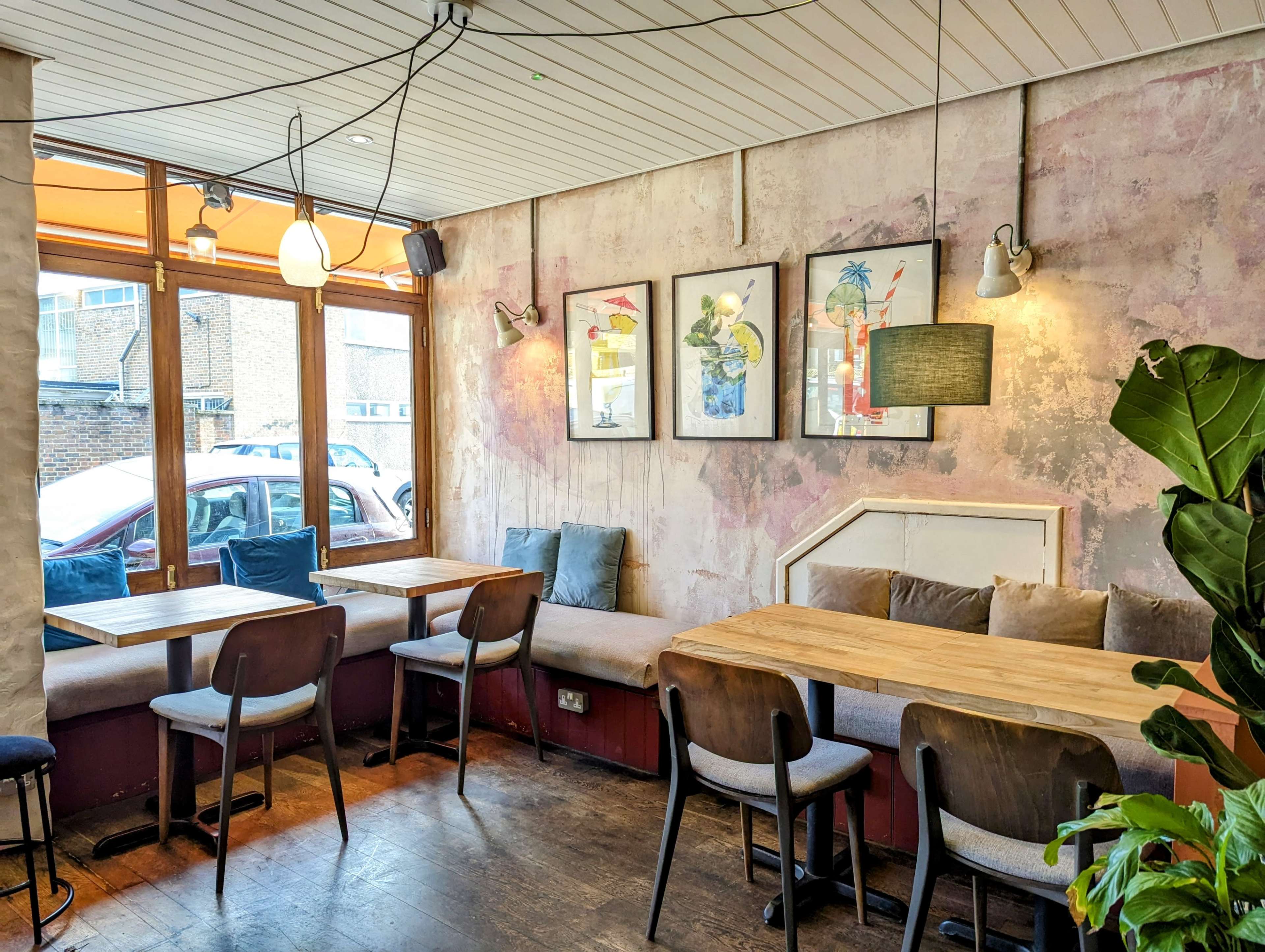 The image shows a cozy café interior with wooden tables, wooden chairs, and framed artwork on the walls, illuminated by hanging lights.