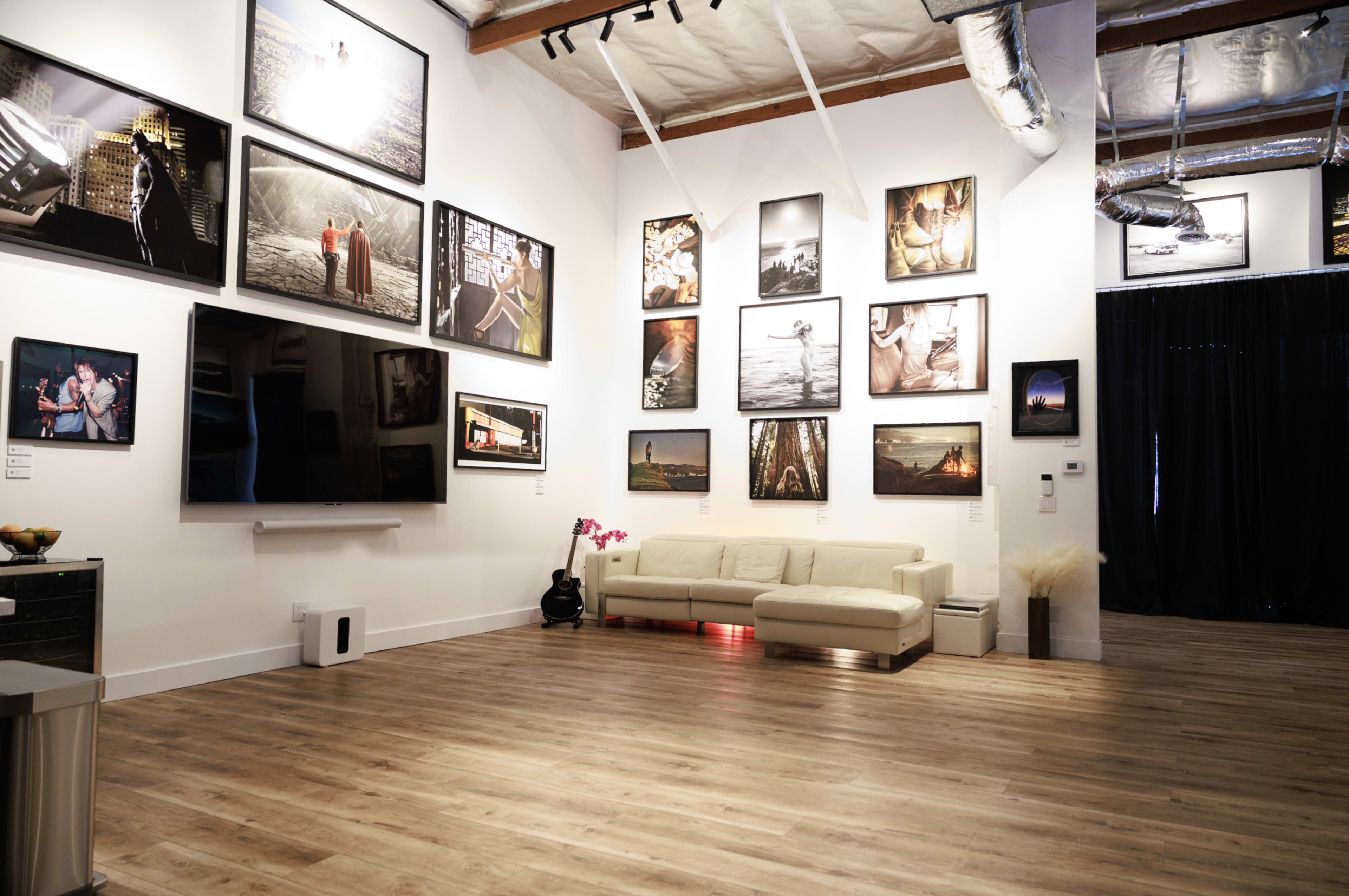 The image features a spacious gallery with a white wall displaying various photographs, a large television, and a light-colored sofa.