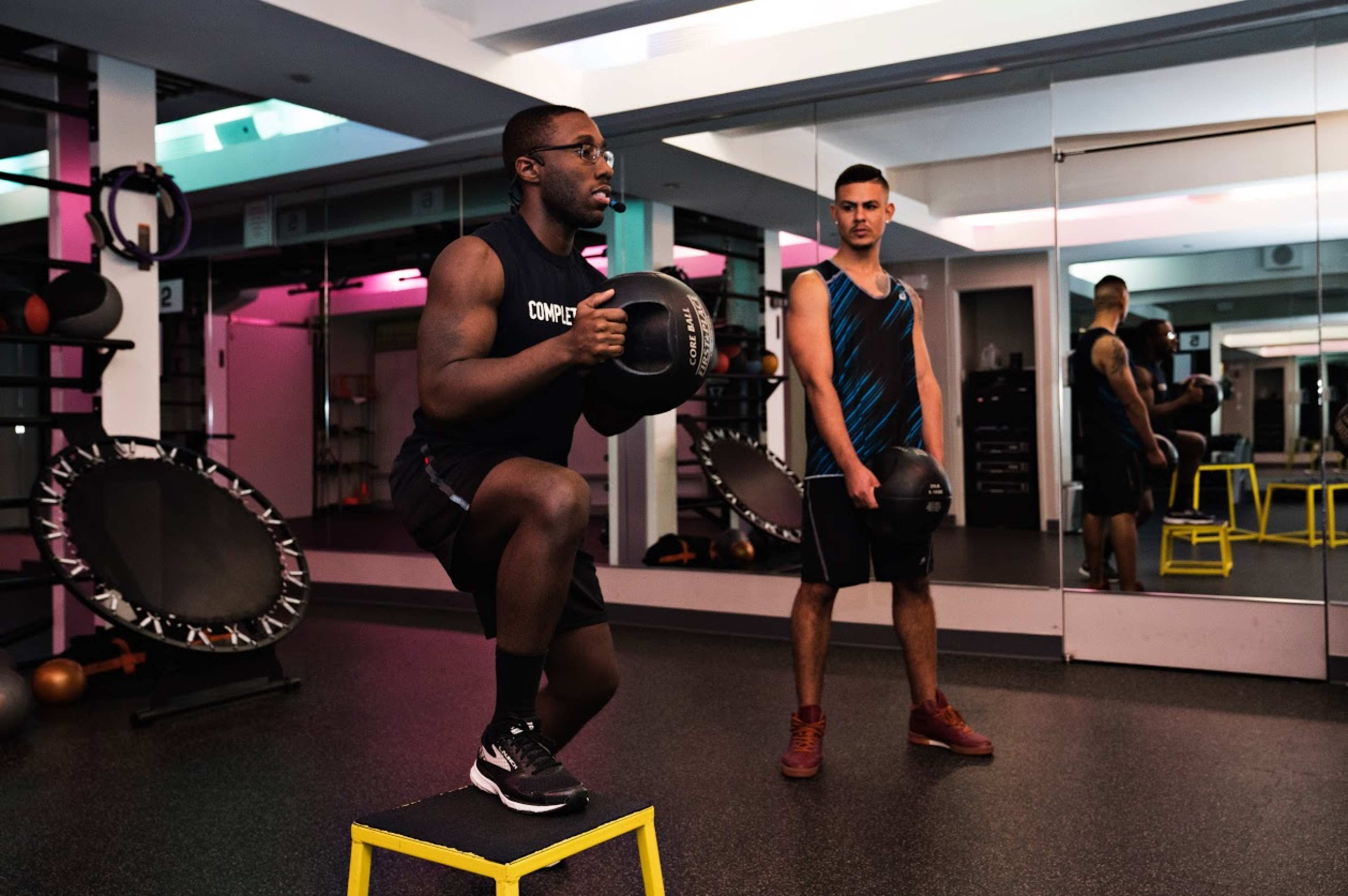 Two men are exercising in a gym, one performing a squat on a platform while holding a dumbbell, and the other observing nearby.