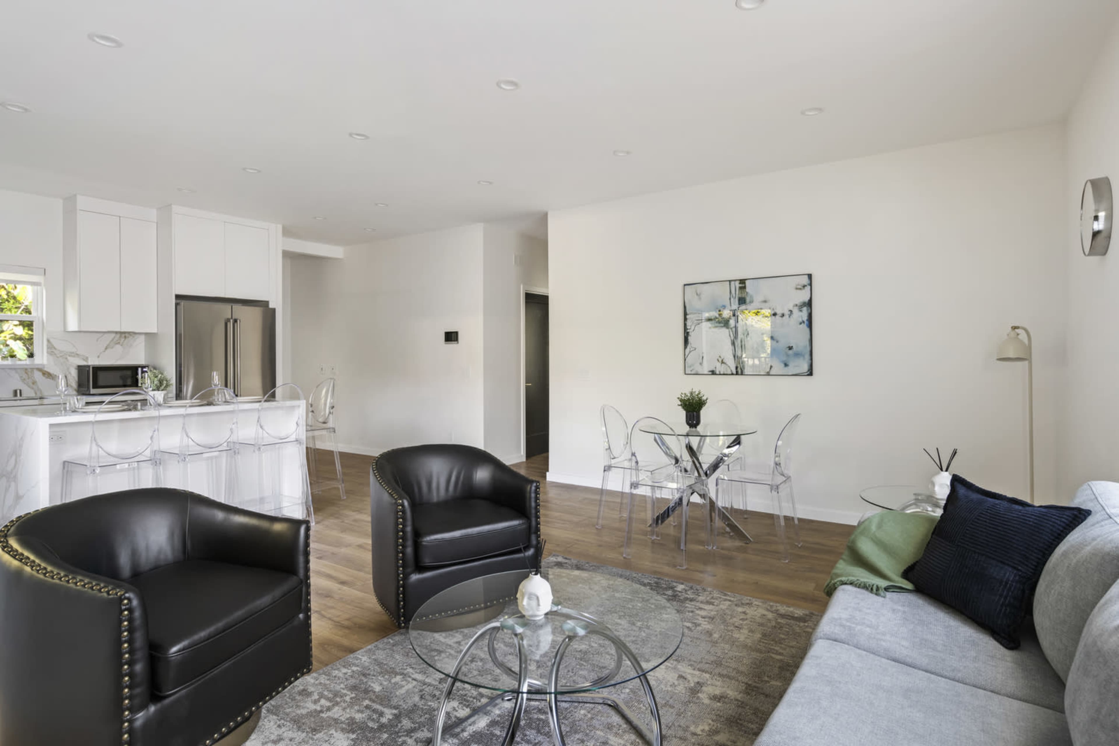 The image shows a modern living area featuring a gray sofa, two black chairs, a glass coffee table, and a dining table with clear chairs, all within a well-lit, open space.