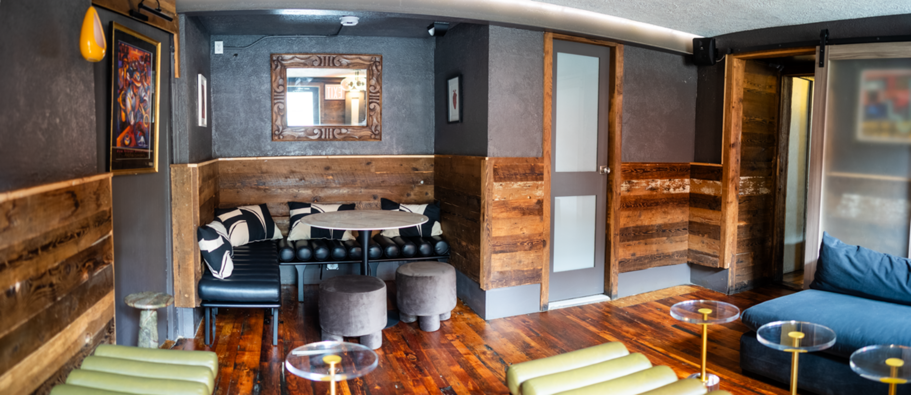 The image shows a small, modern lounge area with wooden accents, a round table, and various seating options, including a black leather couch and two green chairs.