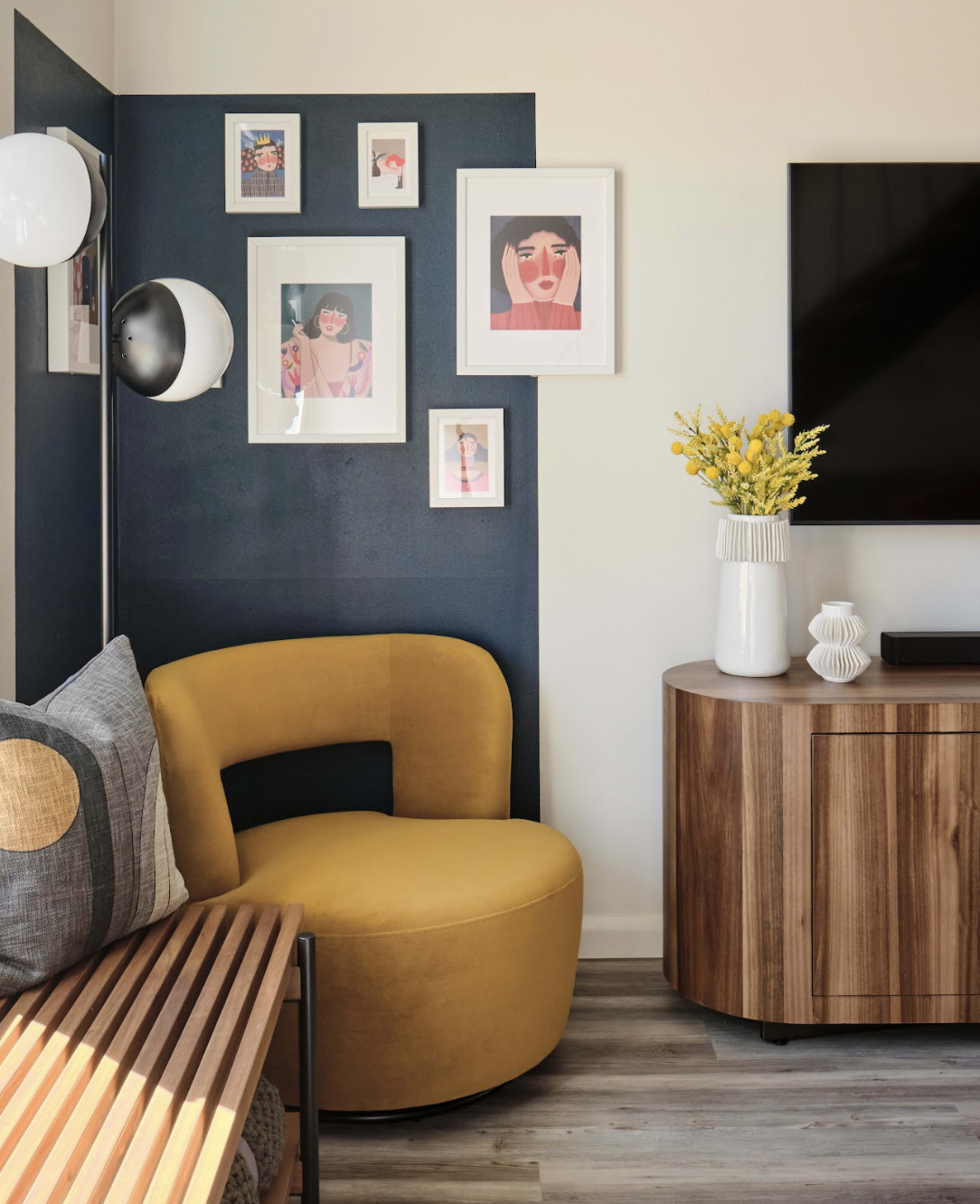 The image depicts a modern living room corner featuring a yellow curved chair, a wooden bench, framed art on the wall, a television, and a decorative vase with flowers on a wooden side table.