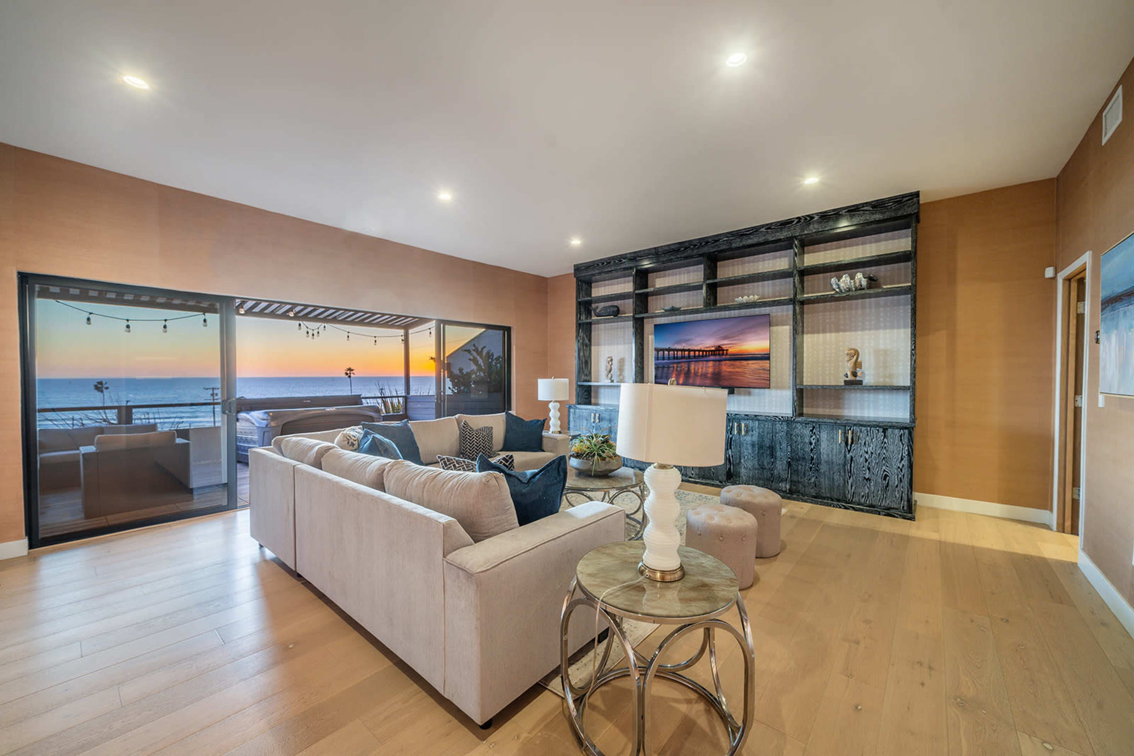 The living room features a sofa facing a mounted television, with large windows showing a view of the ocean at sunset.