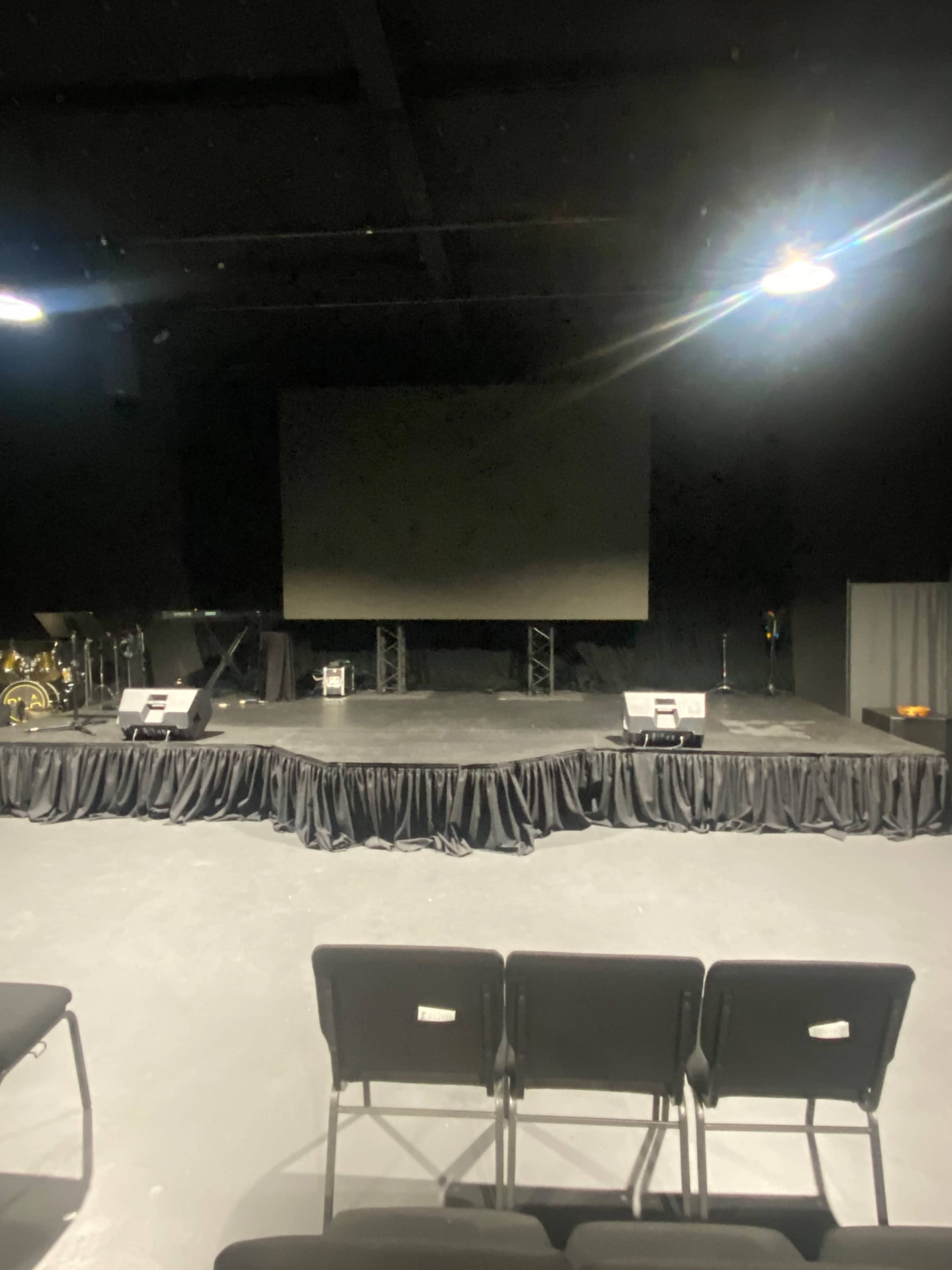 A stage setup in a dimly lit venue with a large screen, speakers, and chairs facing the stage.