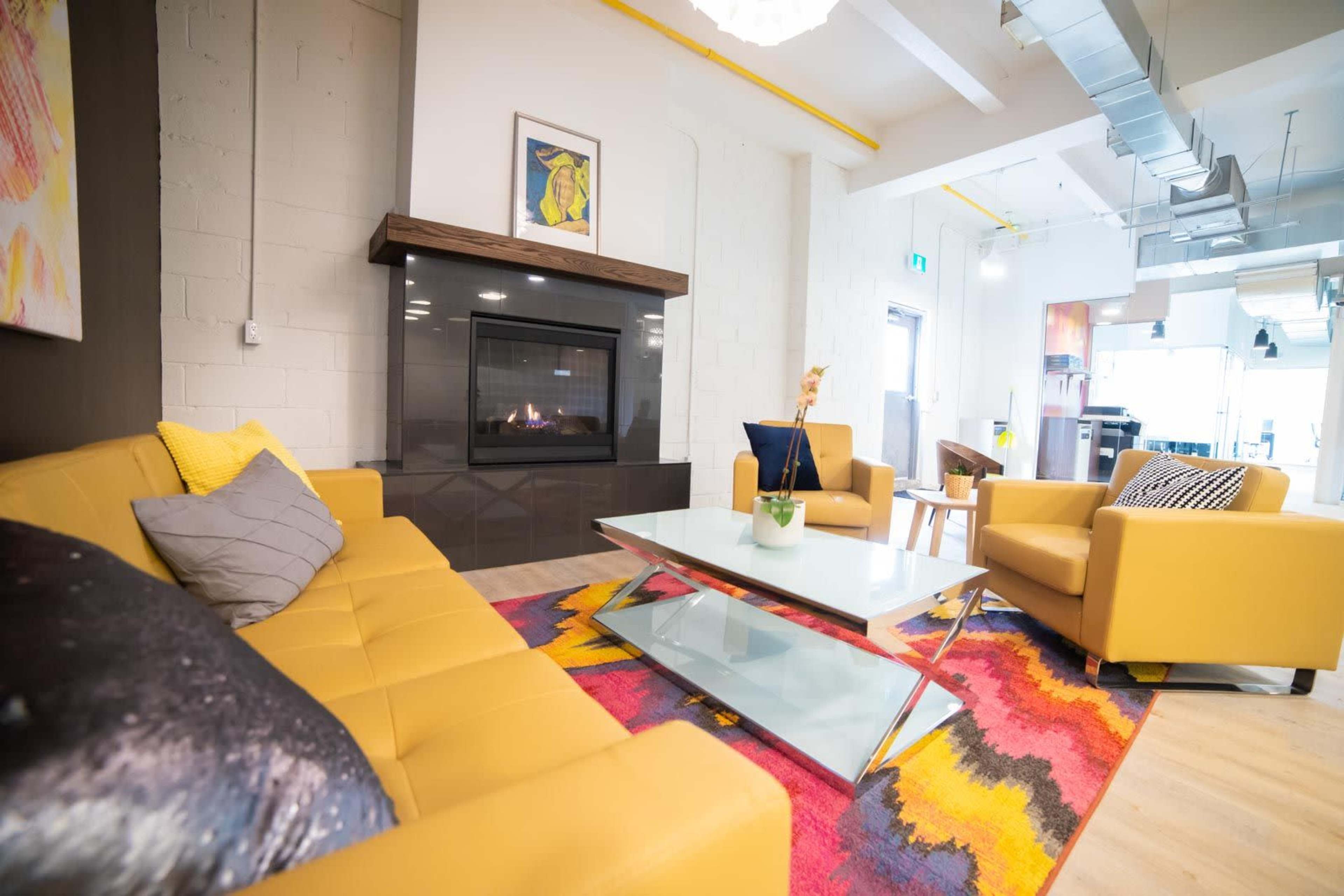 A modern living area features a yellow sectional sofa, a glass coffee table, and a fireplace, with a colorful rug and artwork on the walls.