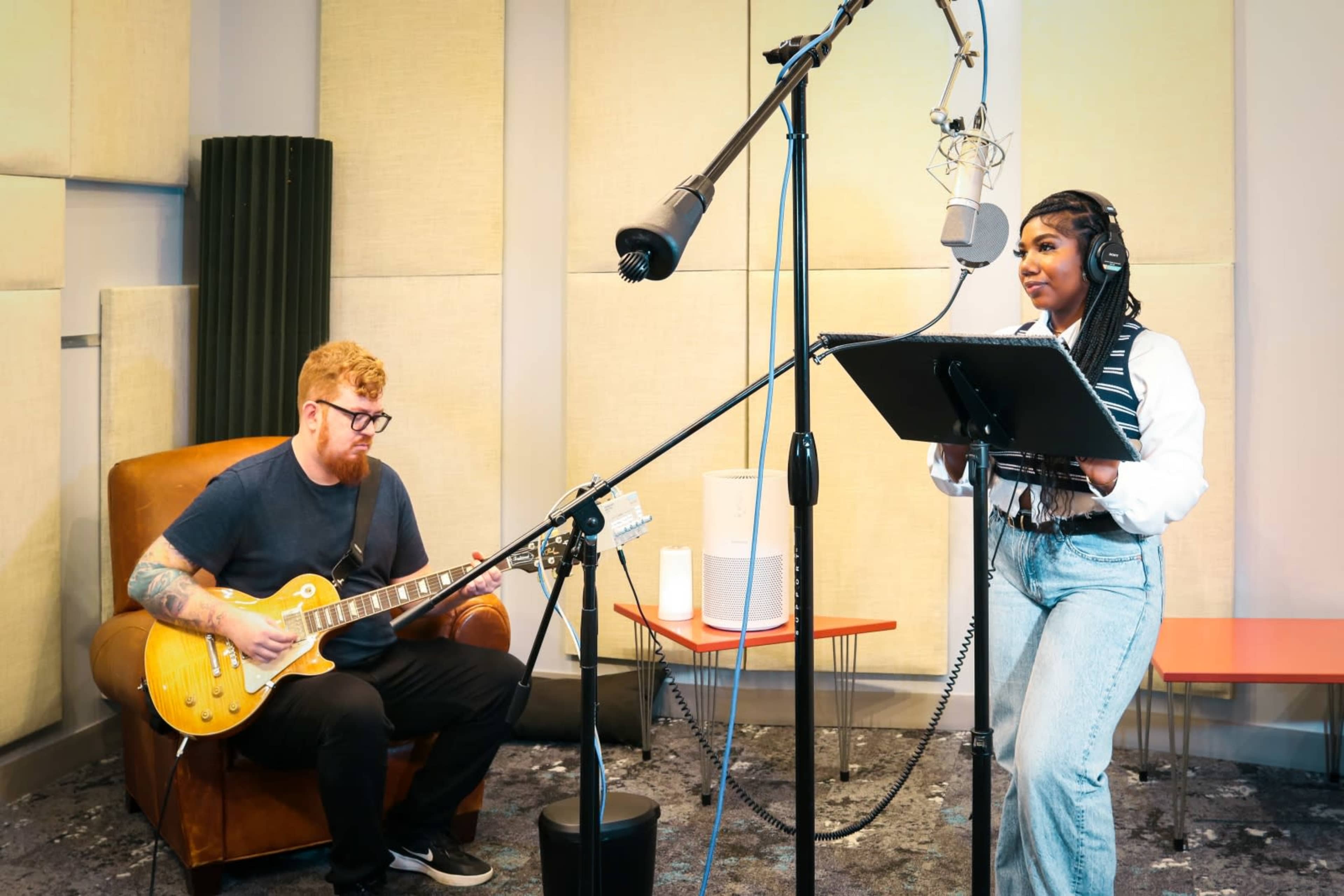 A musician sings into a microphone while another plays guitar in a recording studio.