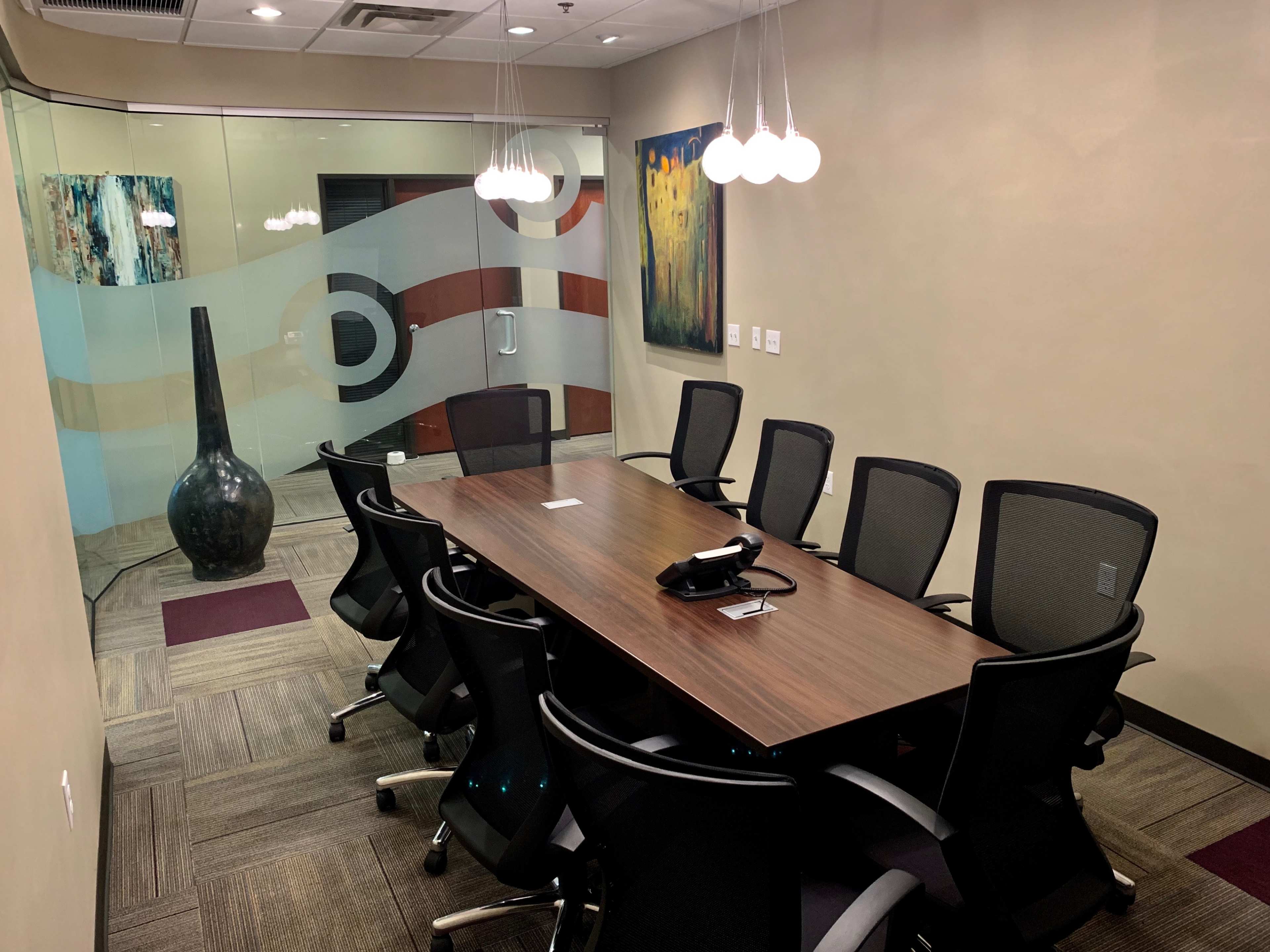 A conference room features a long wooden table surrounded by several ergonomic chairs, with a large black vase and abstract wall art in the background.