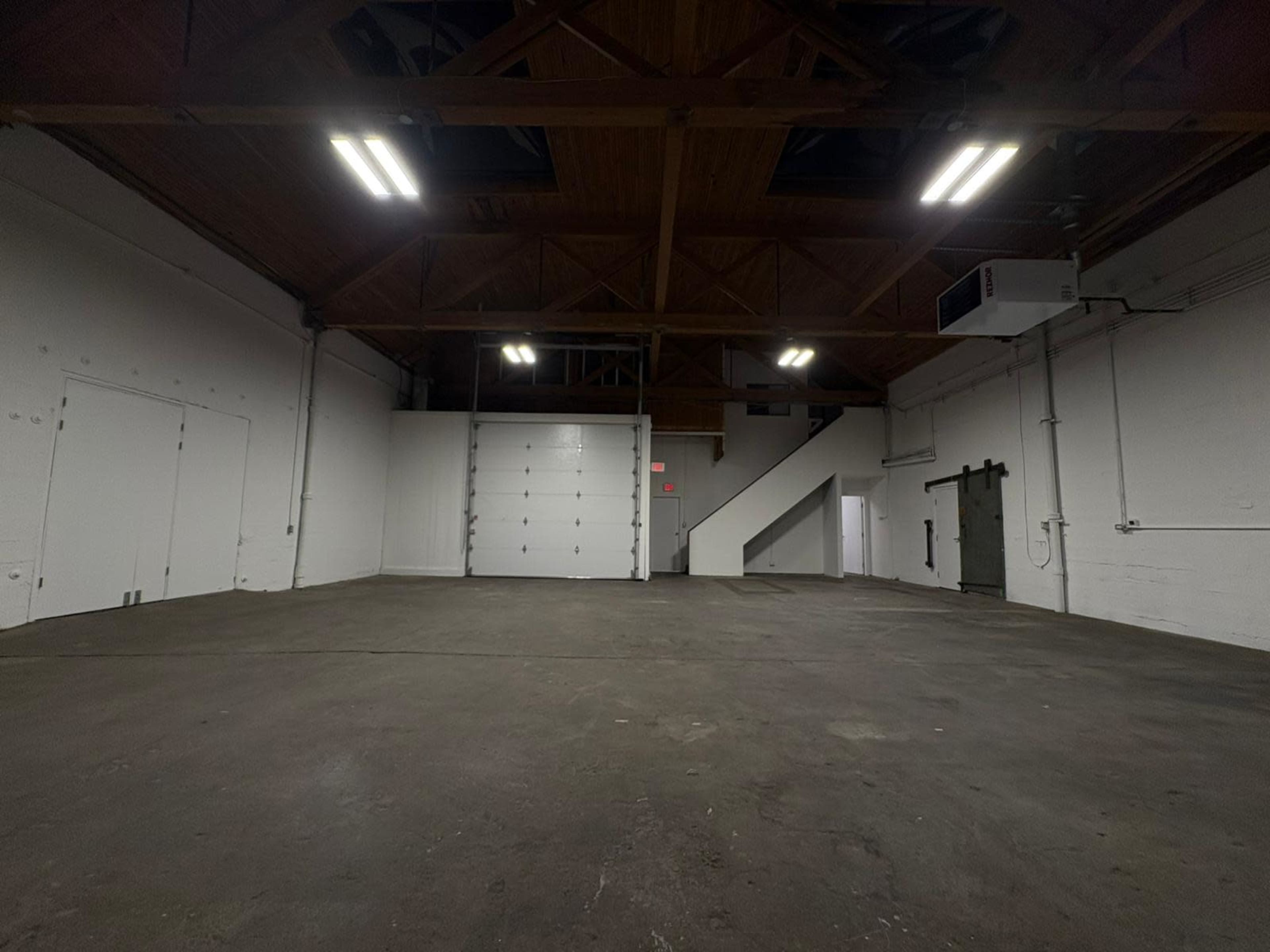 The image shows an empty warehouse interior with a high, wooden-beamed ceiling, concrete floor, and a large garage door.
