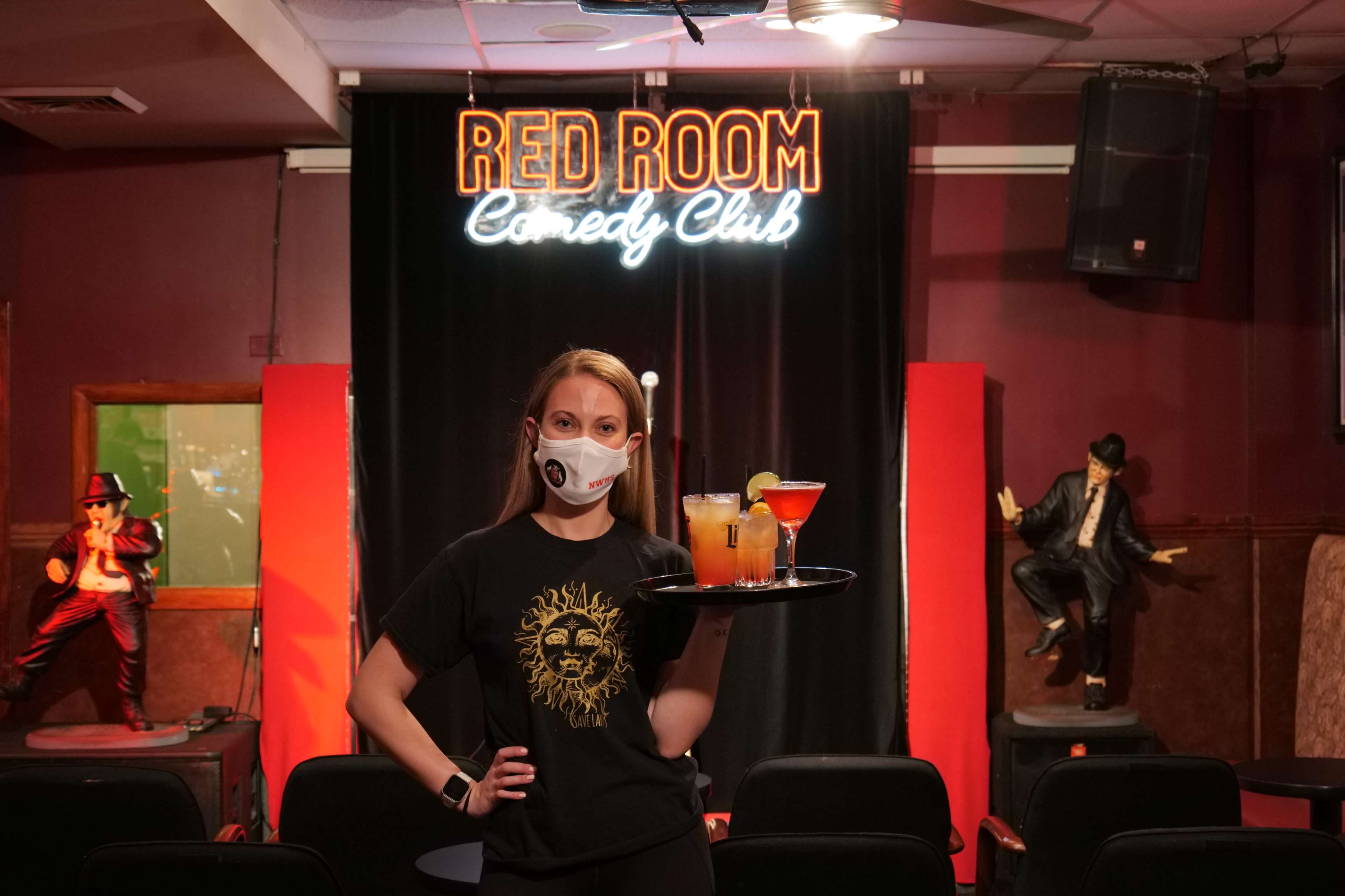 A masked waitress stands in front of the Red Room Comedy Club stage, holding a tray of drinks.