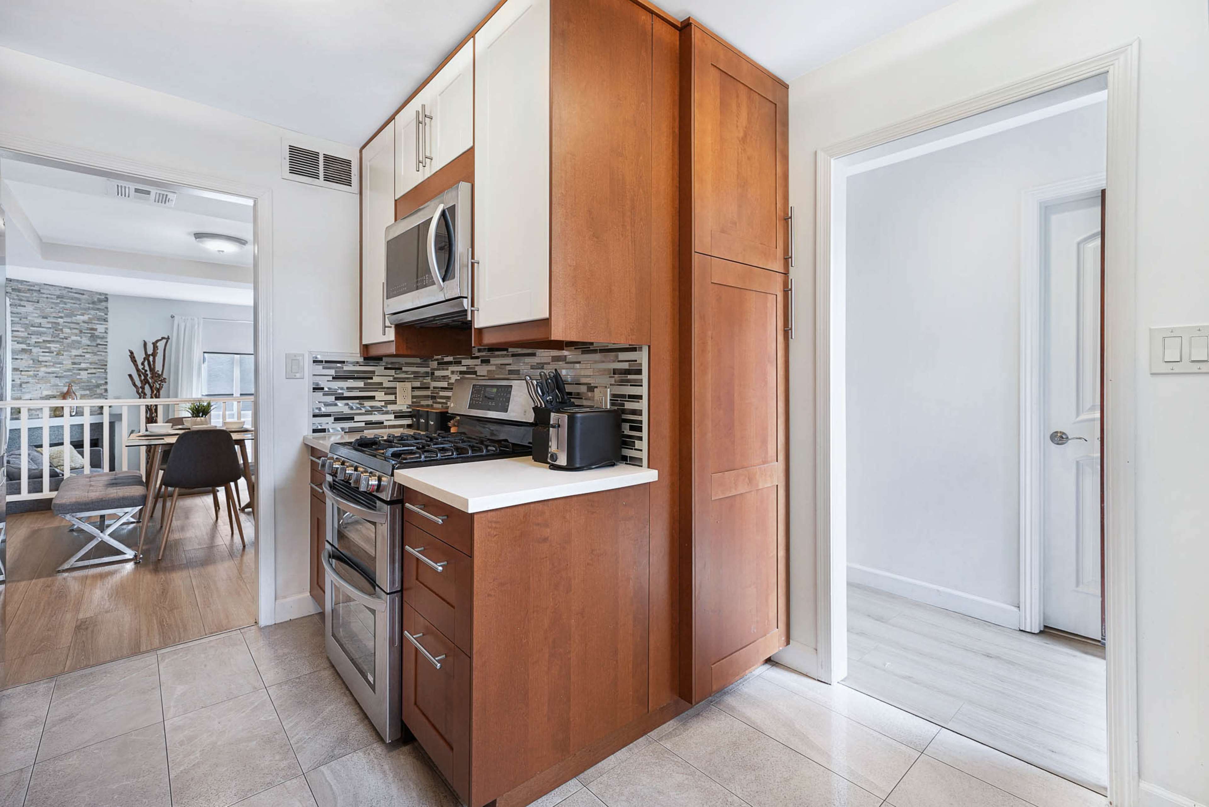 The image shows a compact kitchen featuring wooden cabinets, stainless steel appliances, and a tiled backsplash, with an open doorway leading to another room.
