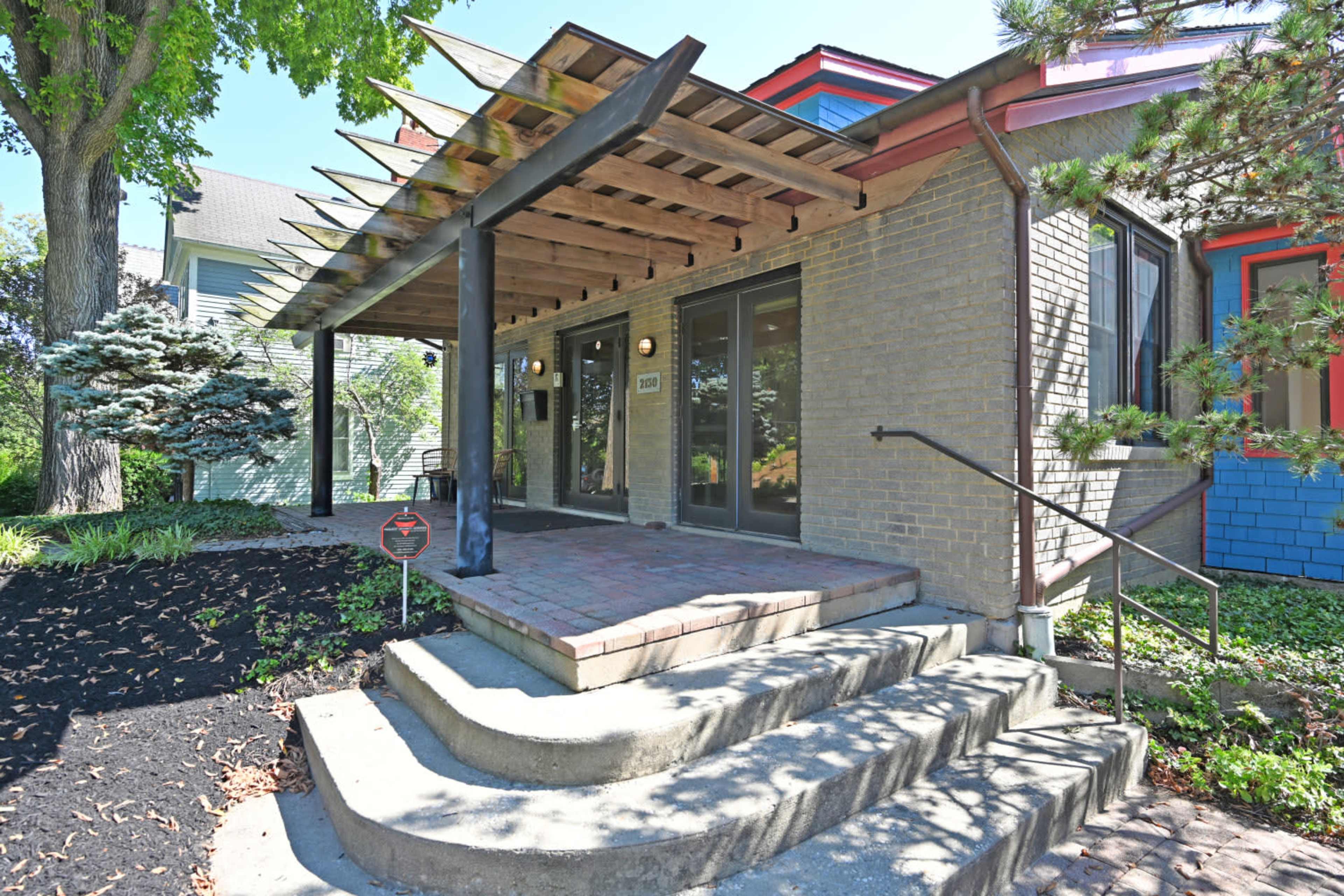 The image shows a building entrance featuring a covered porch with a wooden trellis and steps leading to double glass doors.