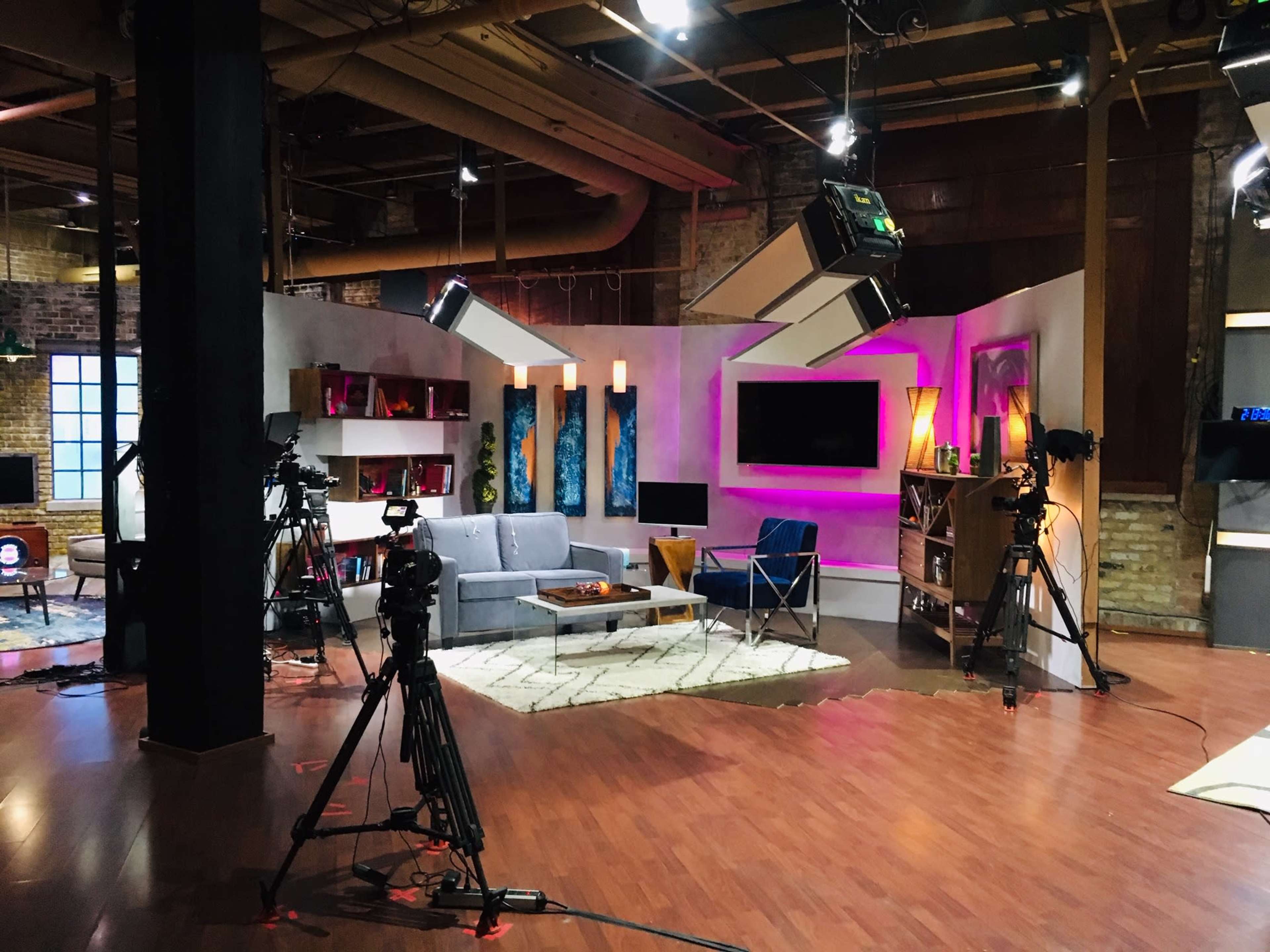 A television studio set with a gray sofa, colorful wall art, and multiple cameras positioned around a brightly lit area.