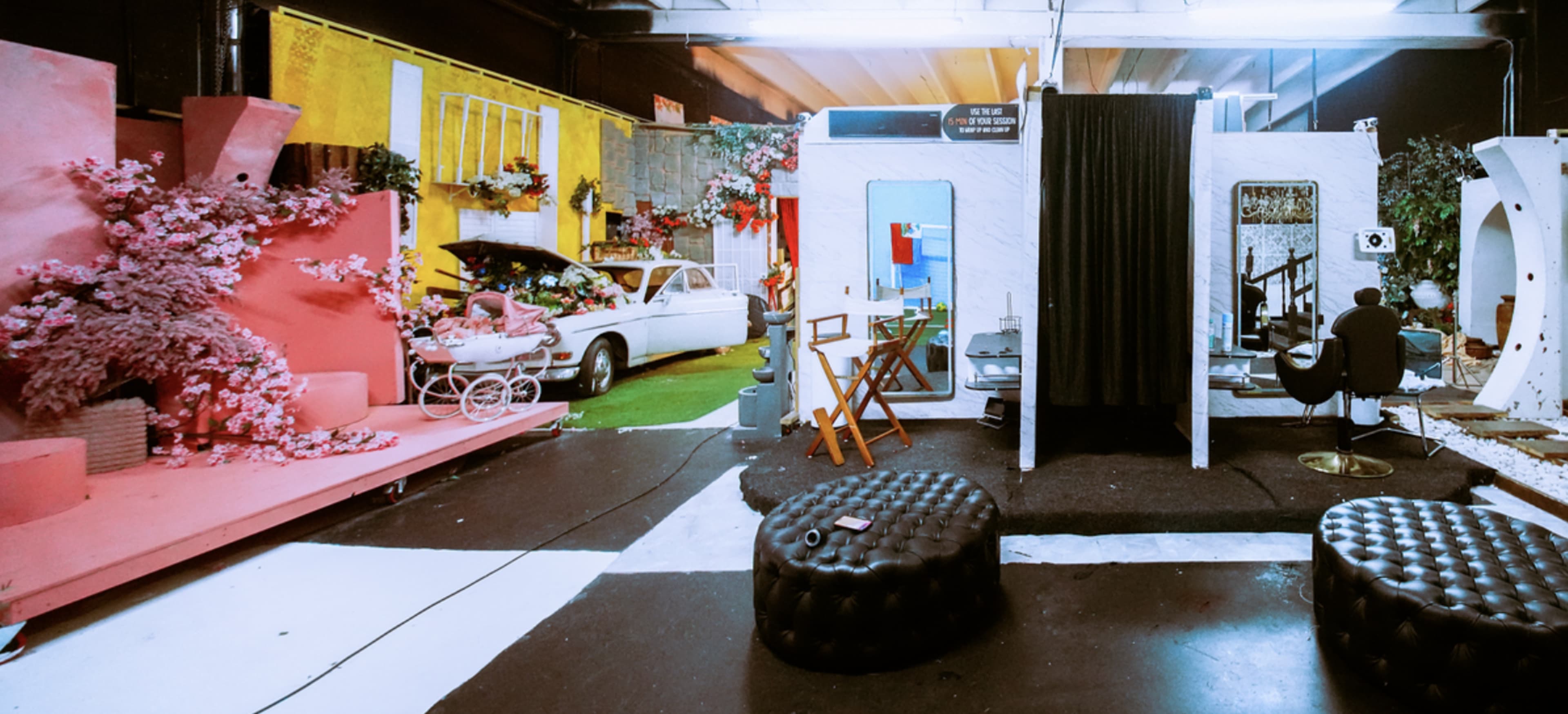 The image shows a vibrant studio space with colorful backdrops, floral arrangements, a vintage car, and various furniture pieces set up for photography or filming.