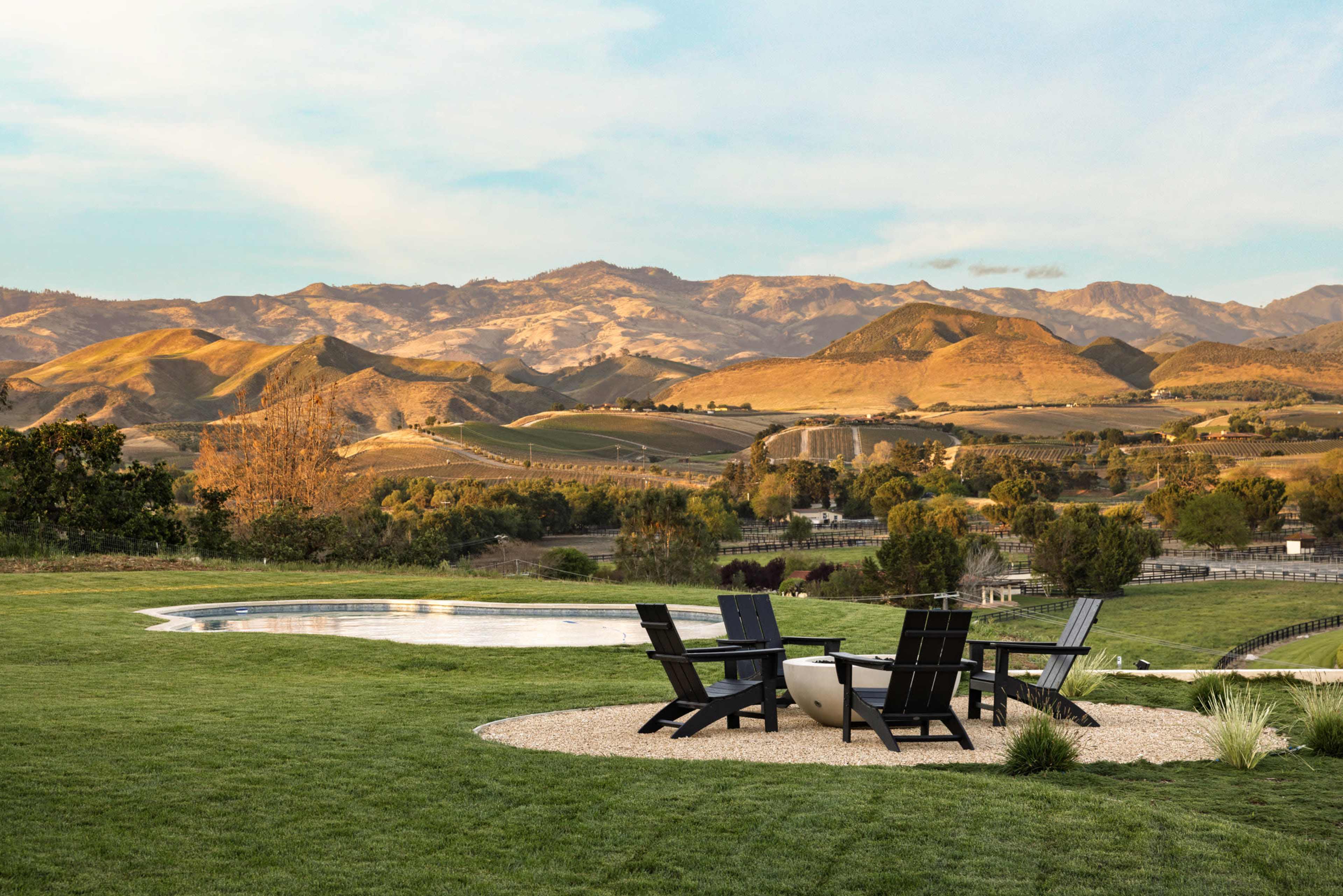 A seating area with black chairs encircles a small fire pit in a grassy landscape, overlooking rolling hills and mountains.