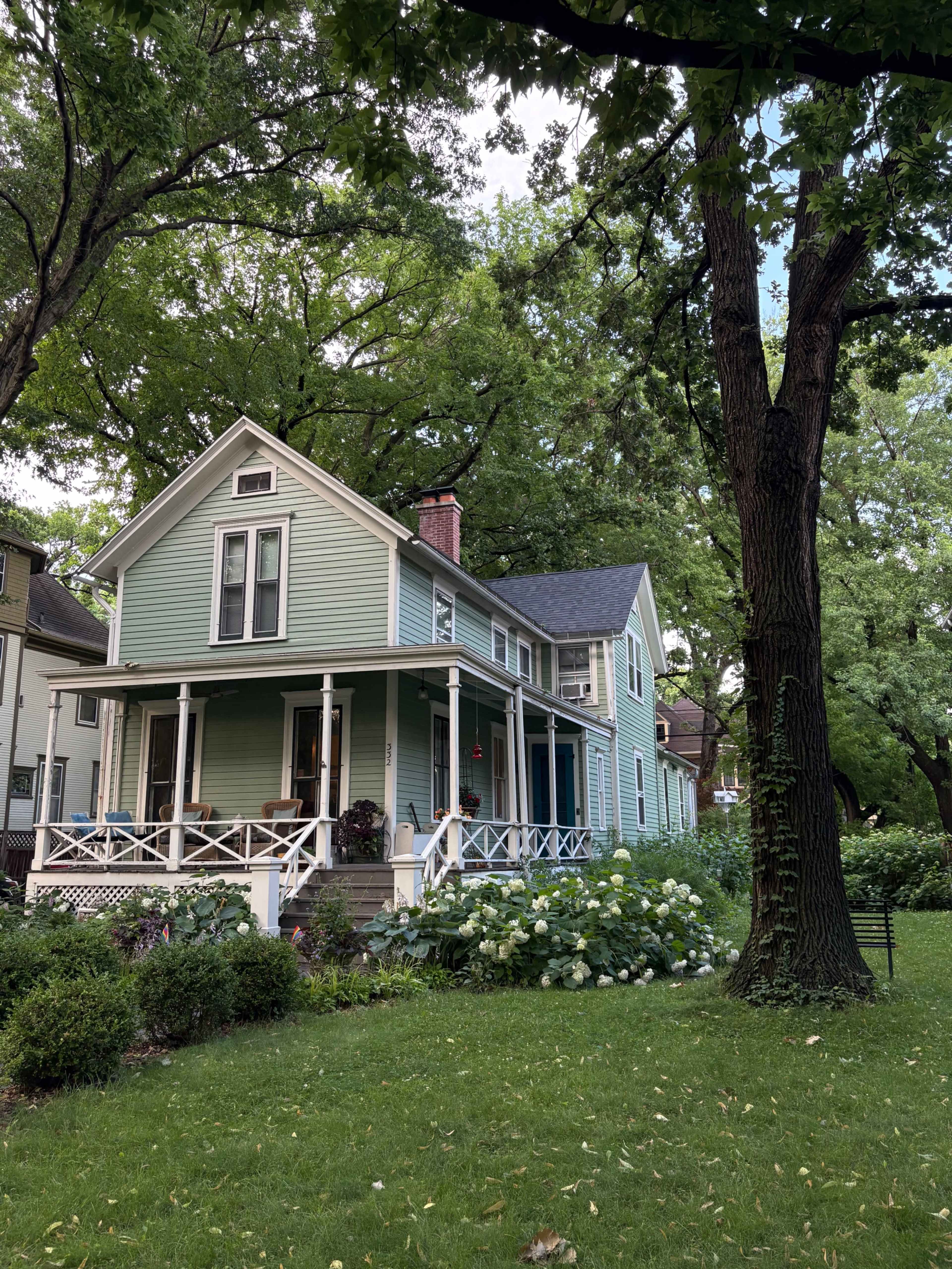 The image shows a green Victorian-style house with a porch, surrounded by trees and shrubs in a well-maintained yard.