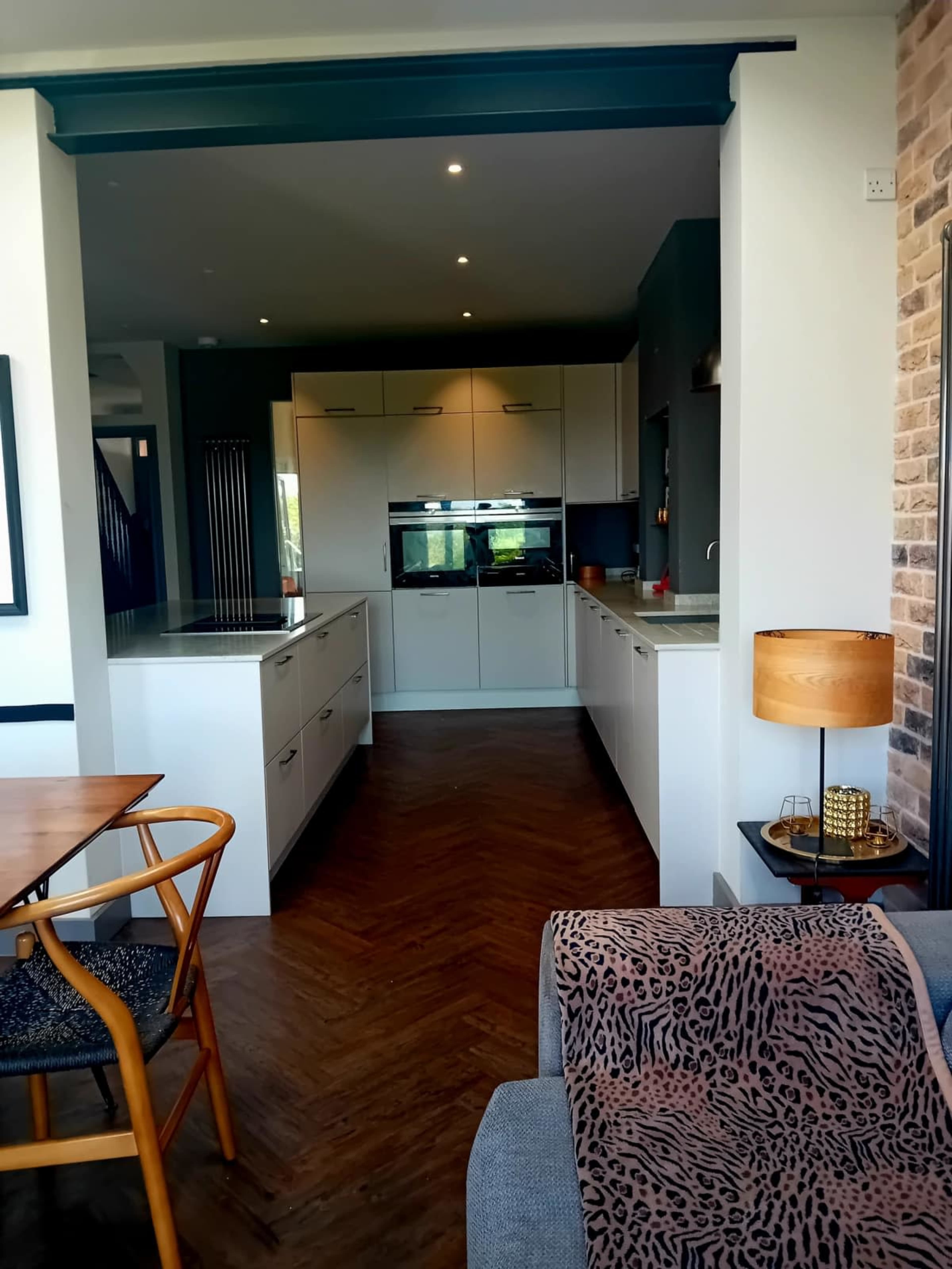 The image shows a modern kitchen with white cabinetry and a sleek countertop, viewed from an open living area that includes a dining table and a decorative lamp.