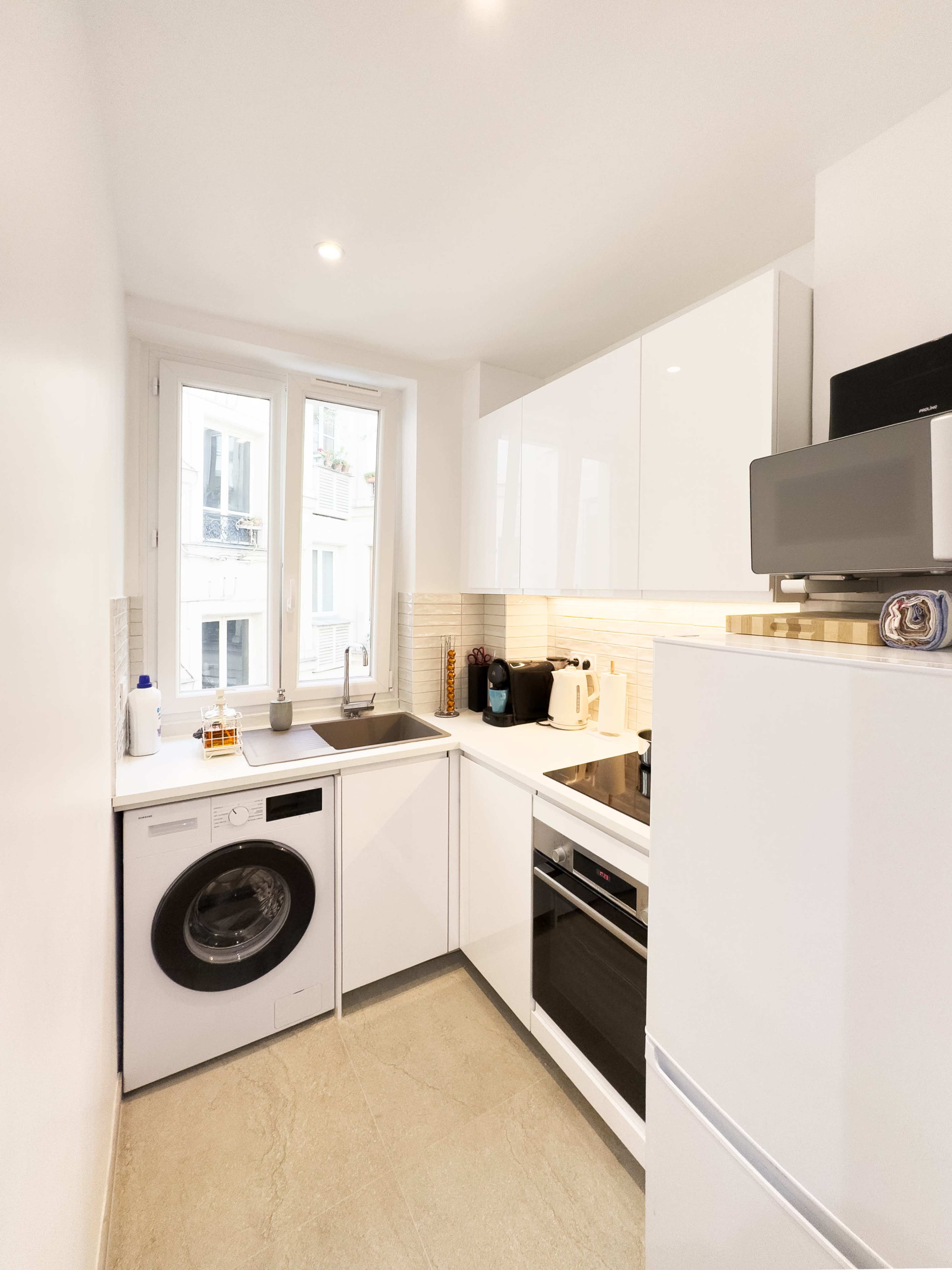 The image shows a small, modern kitchen with white cabinetry, a washing machine, and a window letting in natural light.