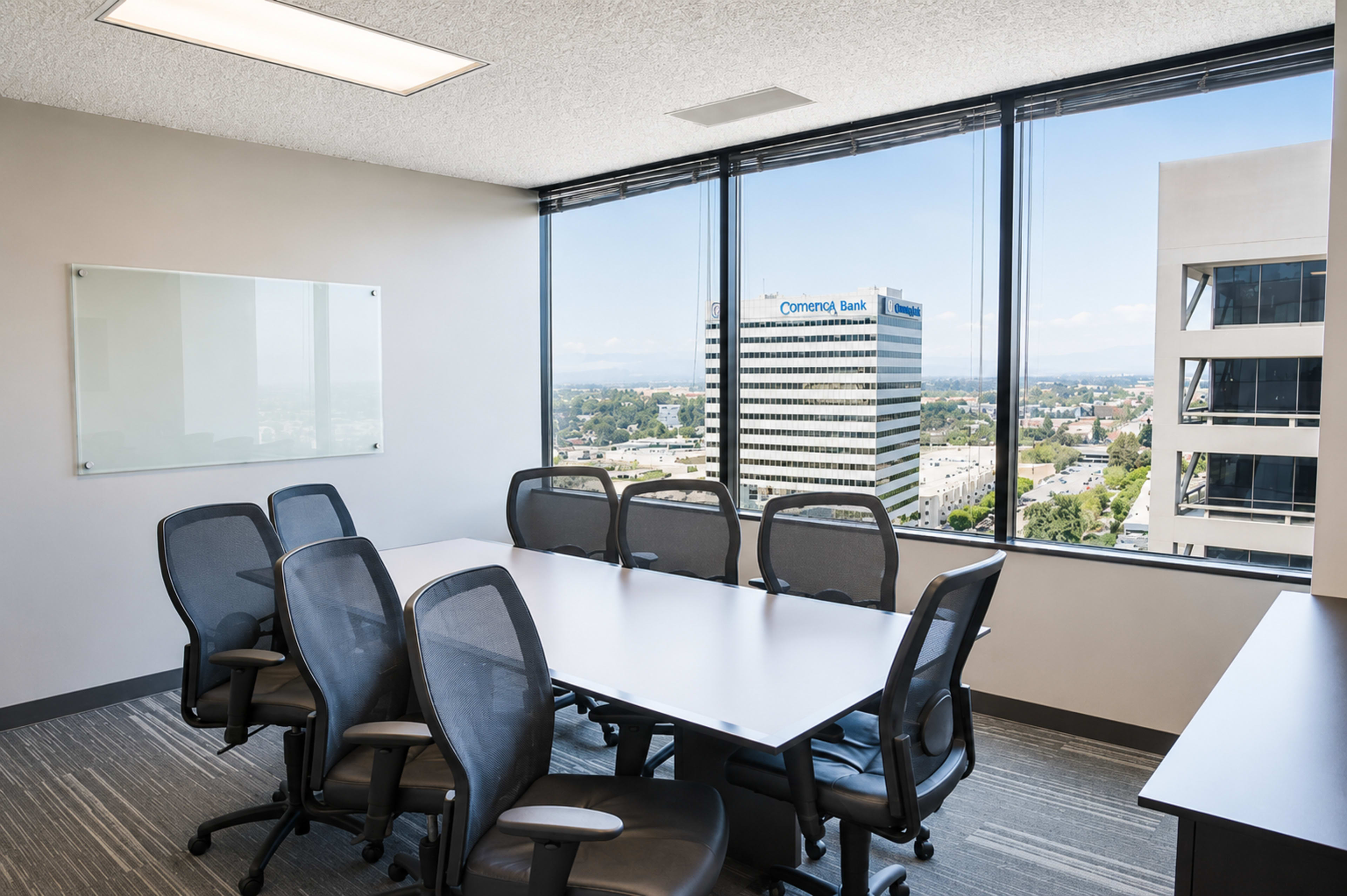 A conference room with a long table and several black mesh chairs features large windows that provide a view of a cityscape and nearby buildings.