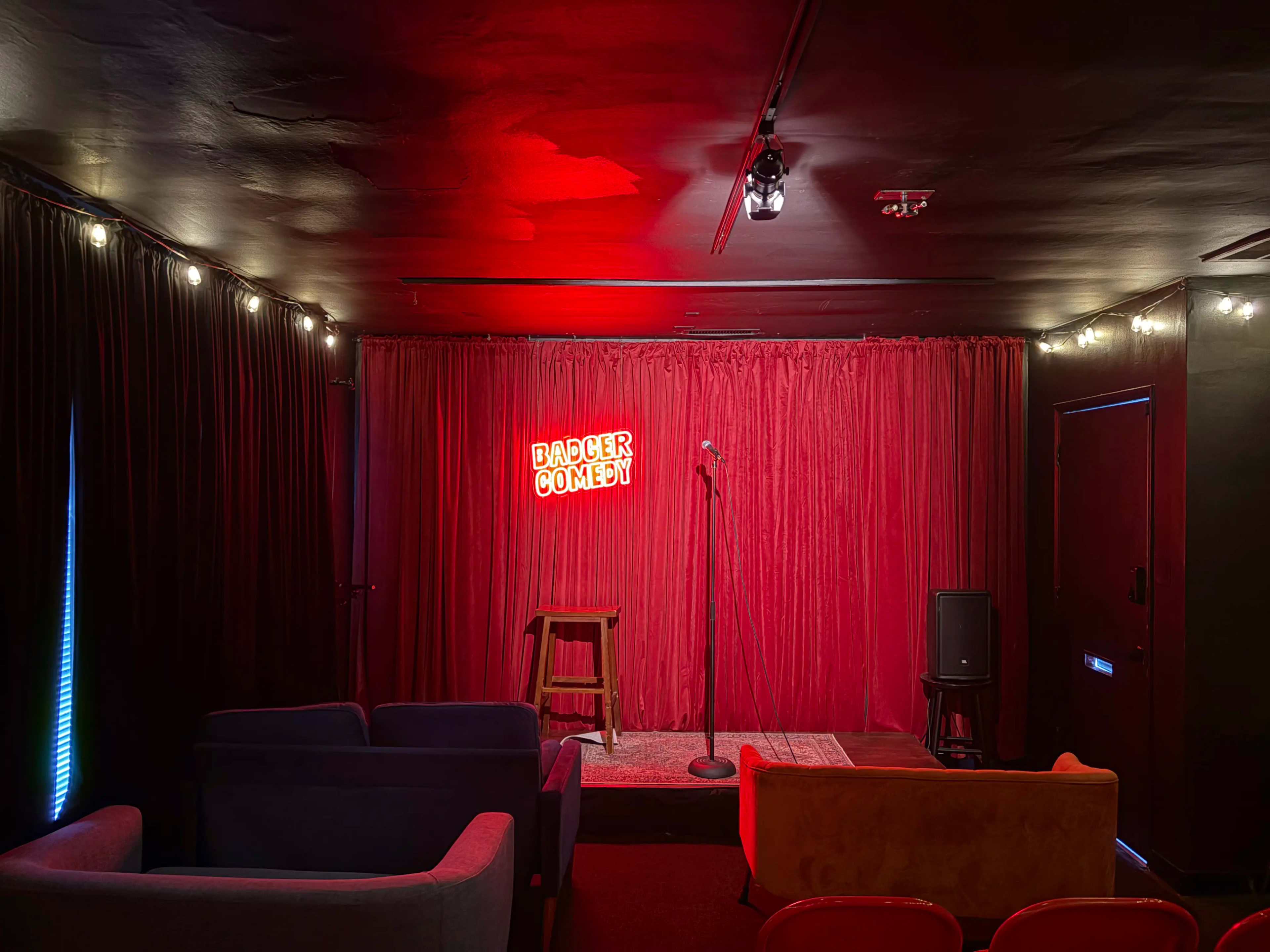 The image shows a small comedy stage with a microphone stand, a bar stool, and a red curtain backdrop, illuminated by a neon sign that reads "Badger Comedy."