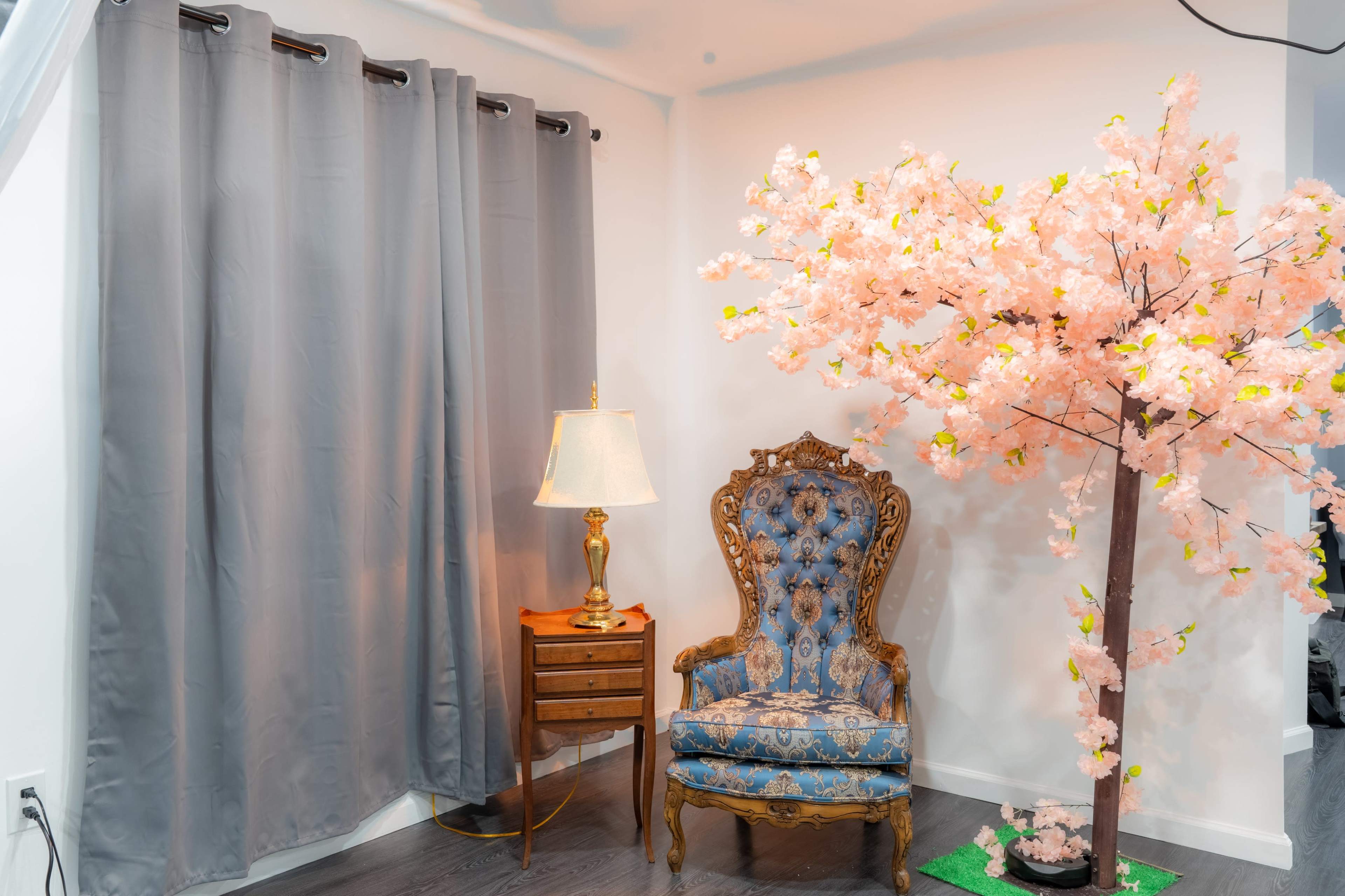 The scene features a blue upholstered chair beside a small wooden table with a lamp, and a decorative pink cherry blossom tree against a backdrop of gray curtains.