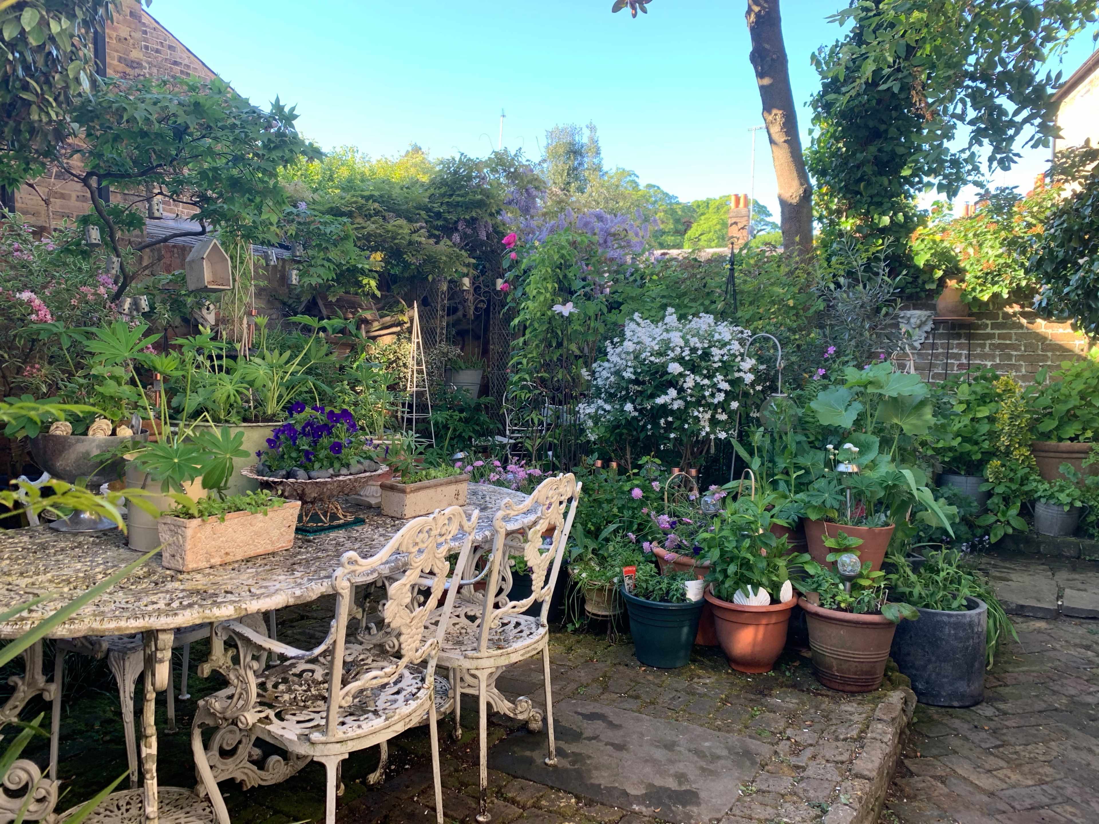 A garden is filled with various potted plants and flowers, with a white wrought-iron table and chairs placed on a stone pathway.