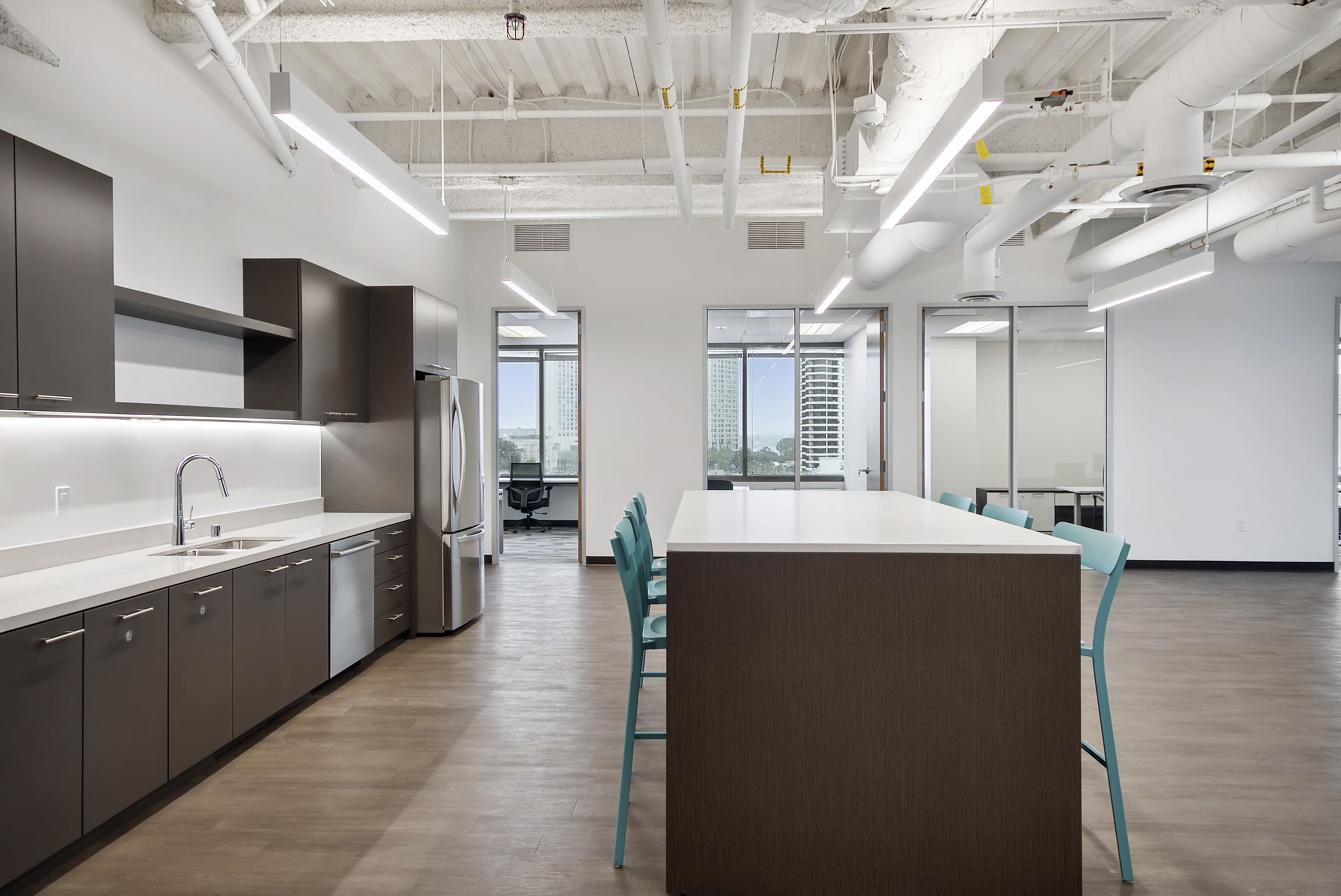 A modern kitchen area in an office featuring stainless steel appliances, dark cabinetry, a large island with seating, and large windows overlooking an outdoor view.