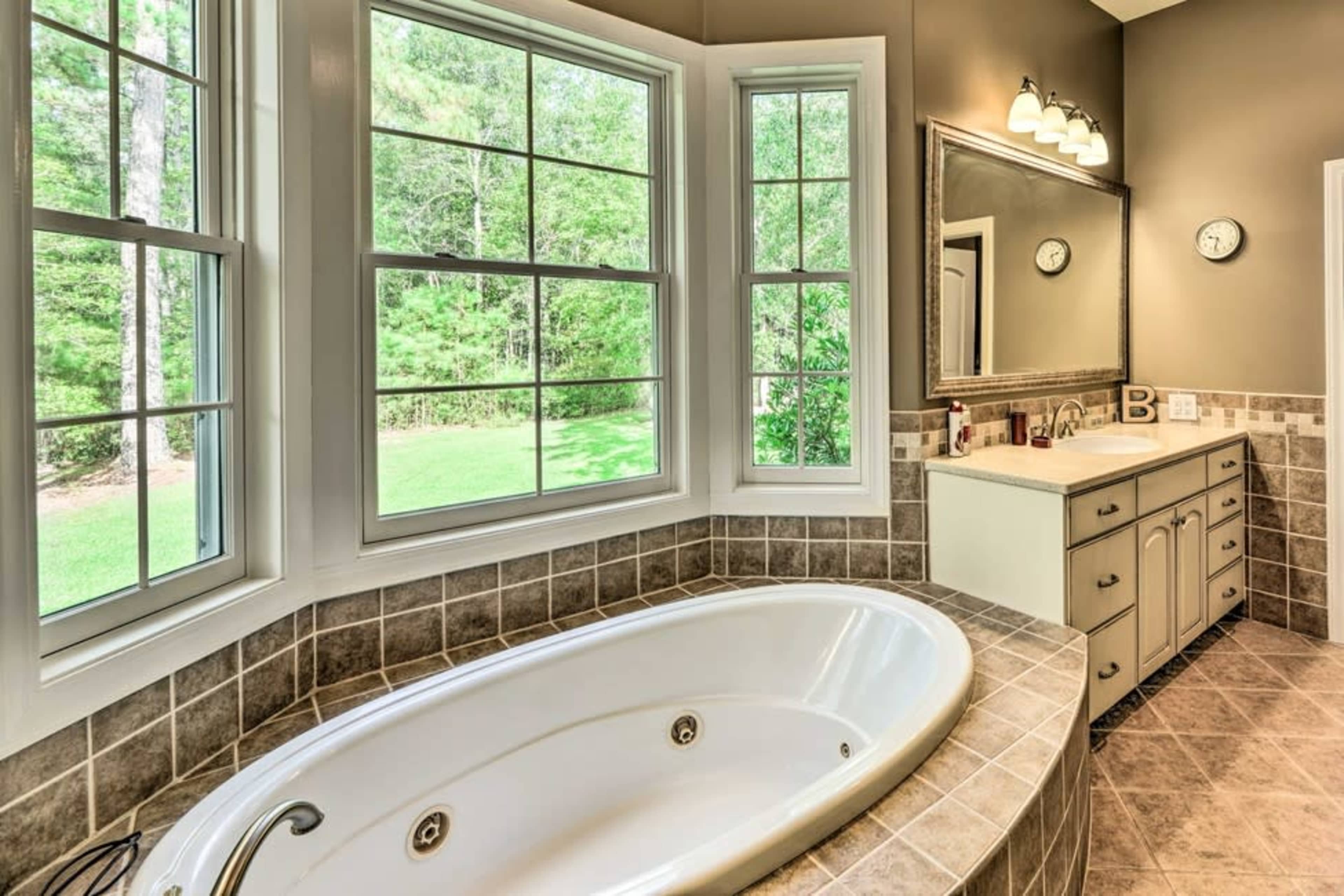 A bathroom featuring a large oval bathtub, a dual sink vanity, large windows overlooking greenery, and tiled walls.