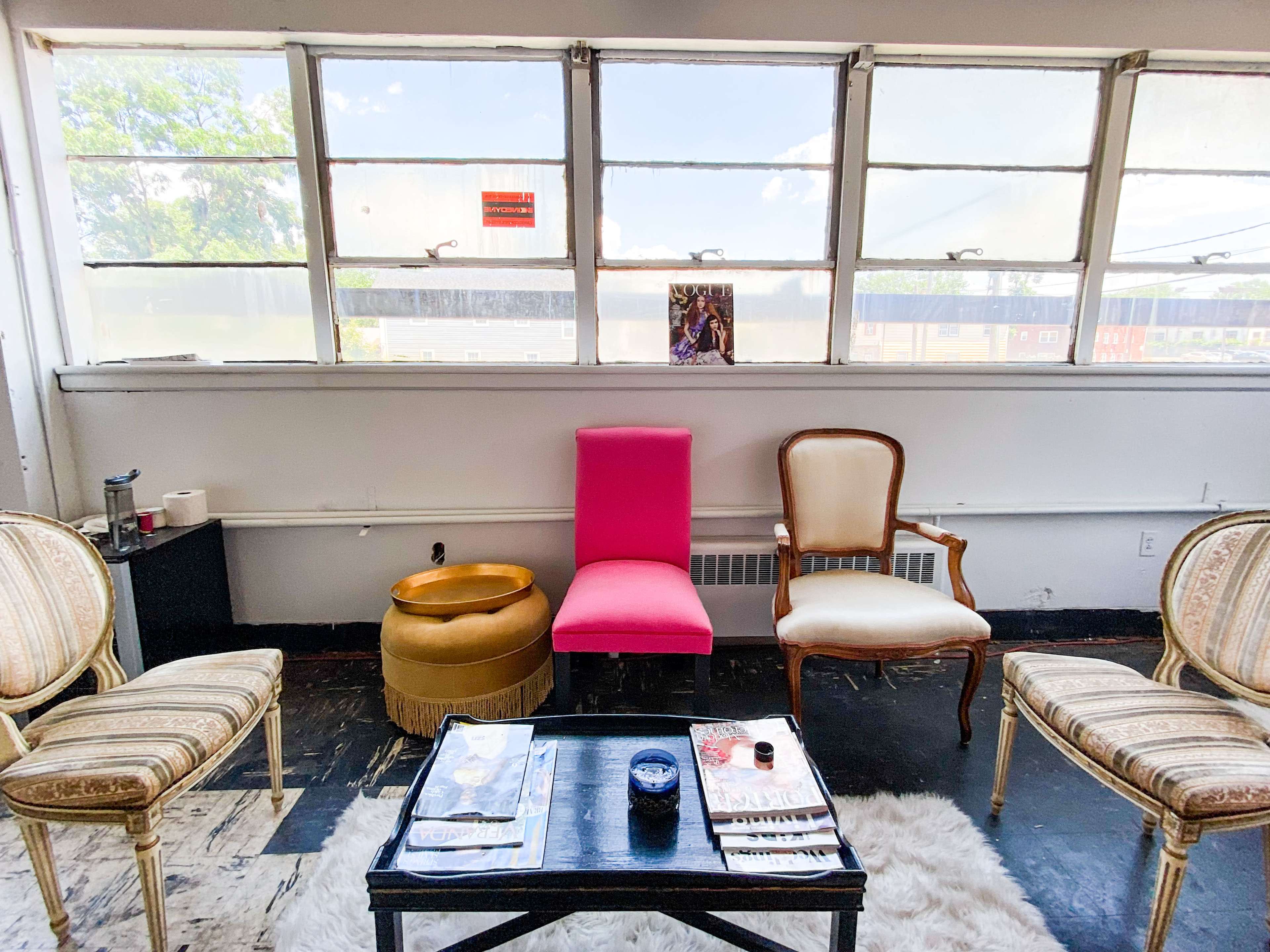 The image shows a bright room with a pink chair, a wooden table displaying magazines, and two patterned chairs facing a large window.