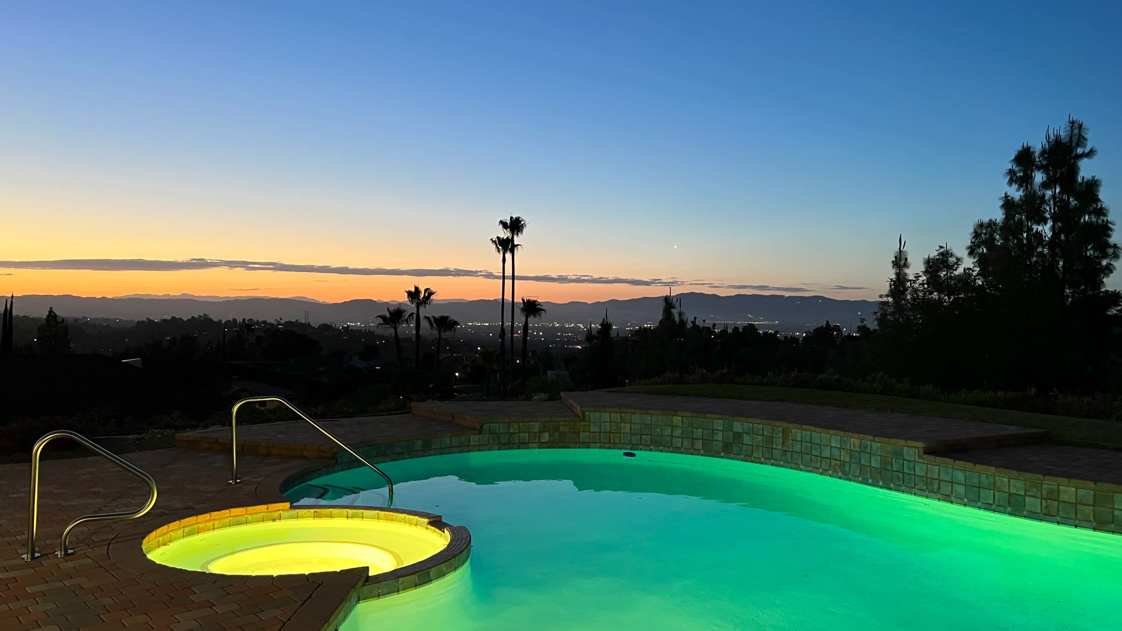 A swimming pool with a built-in hot tub is illuminated by green lighting against a backdrop of a sunset and distant mountains.