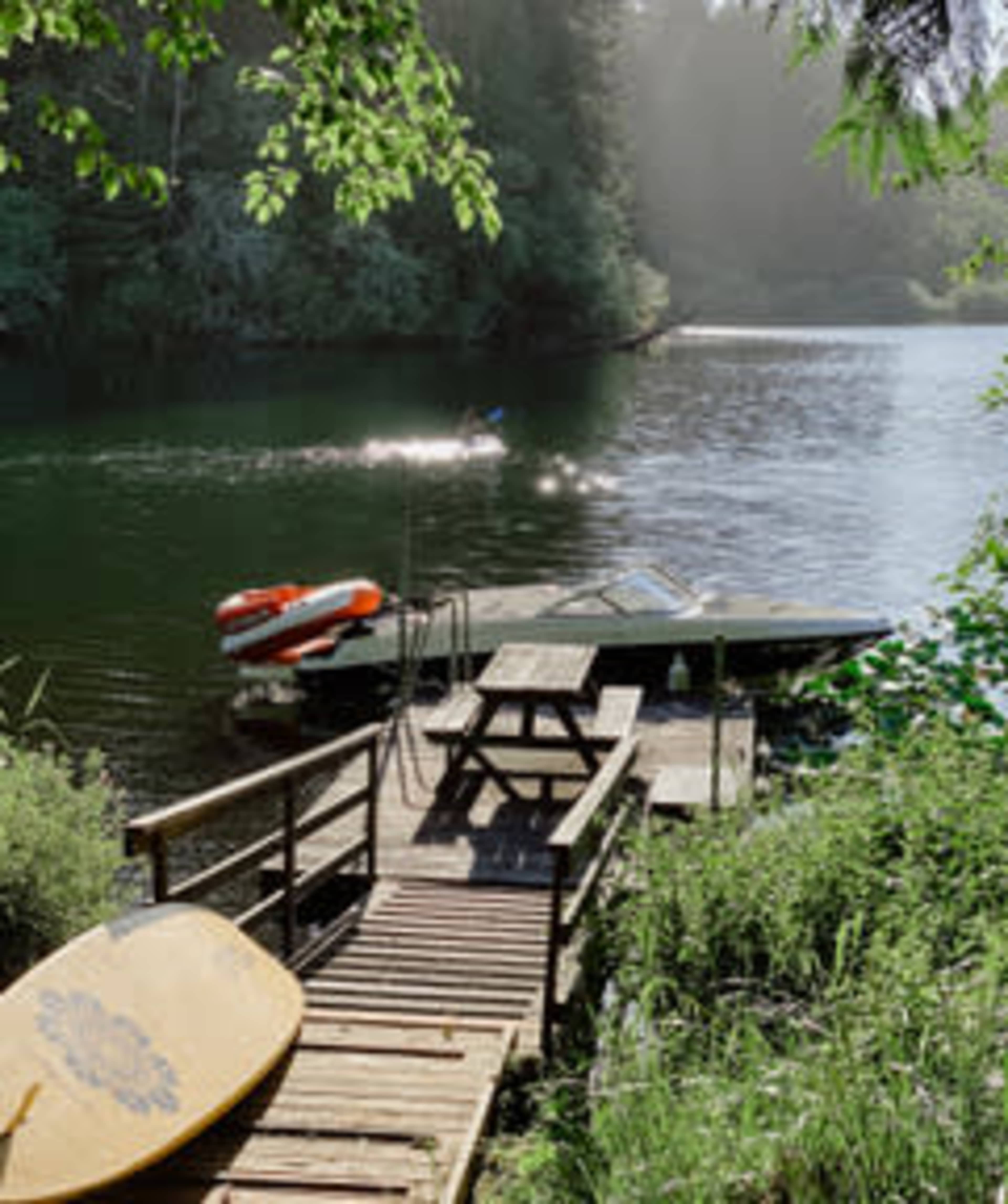 A wooden dock extends over a calm lake where a small boat is moored and a paddleboard rests nearby.