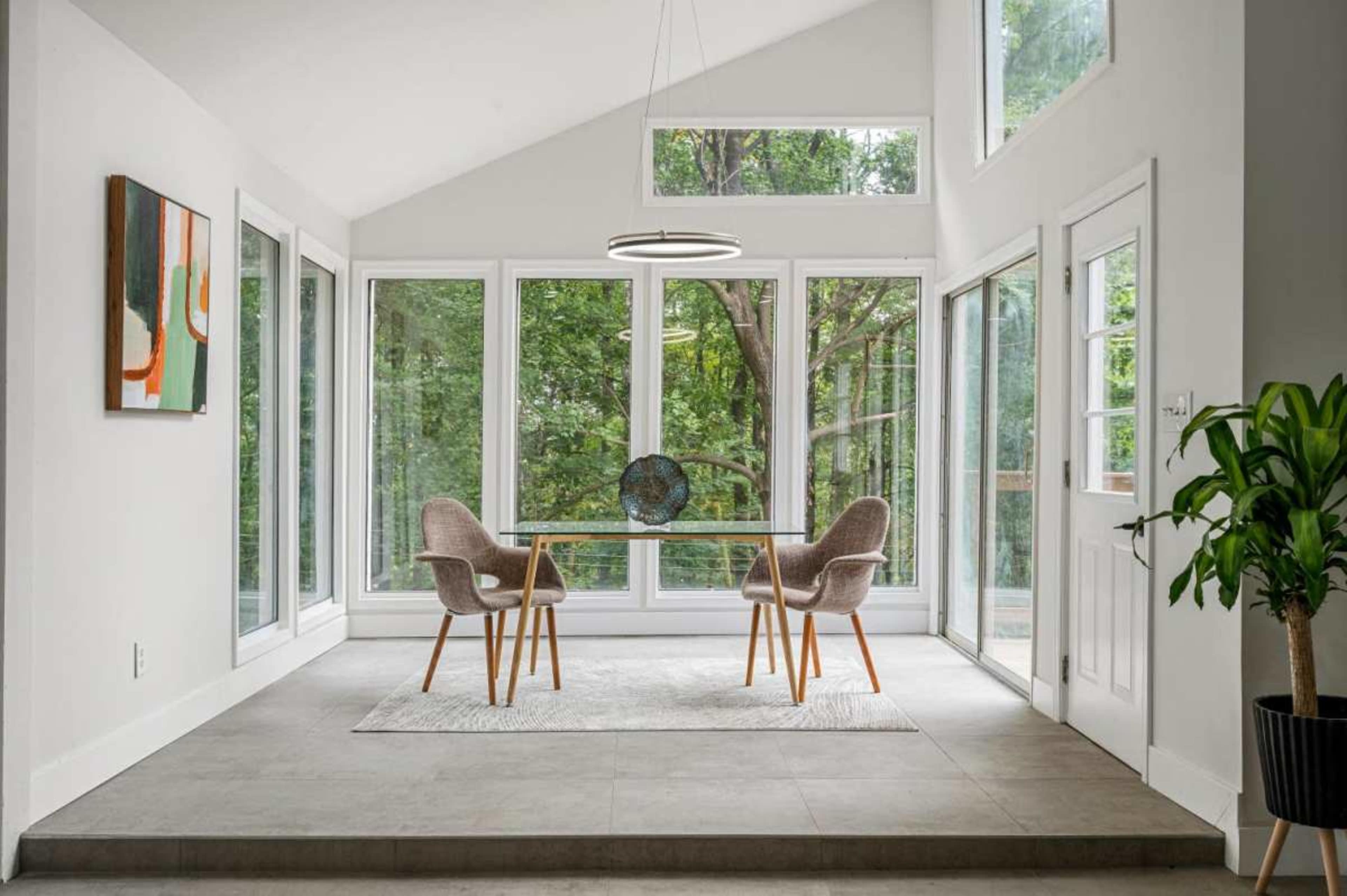 A modern sunlit dining area features a glass table with two chairs, surrounded by large windows overlooking a lush outdoor view.