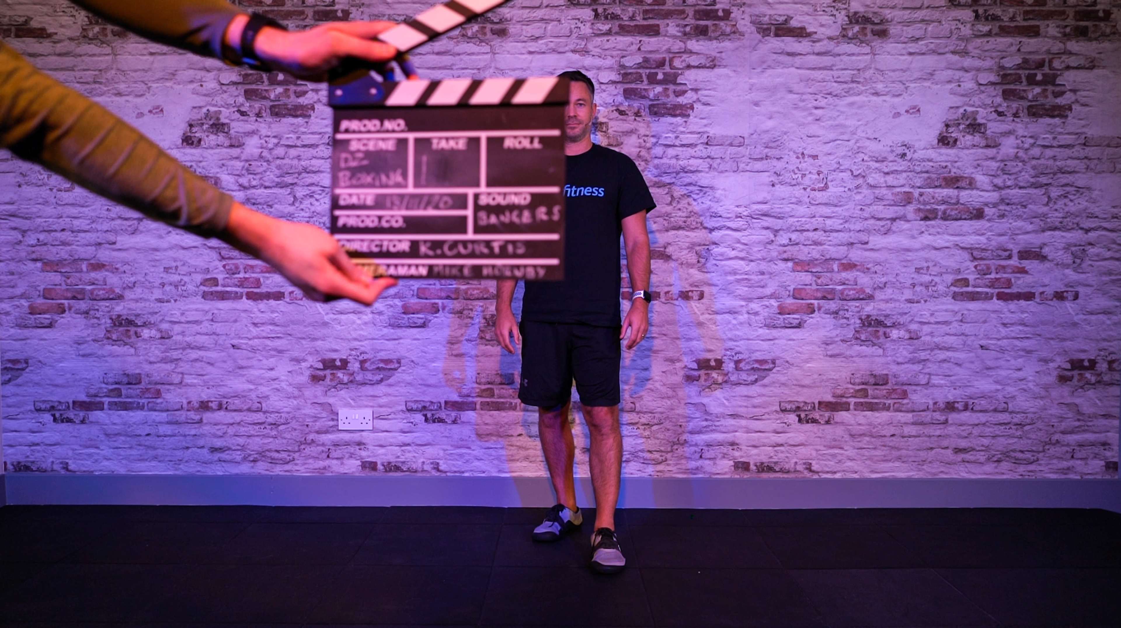 A person stands against a brick wall while another person holds a movie clapperboard in front of them.