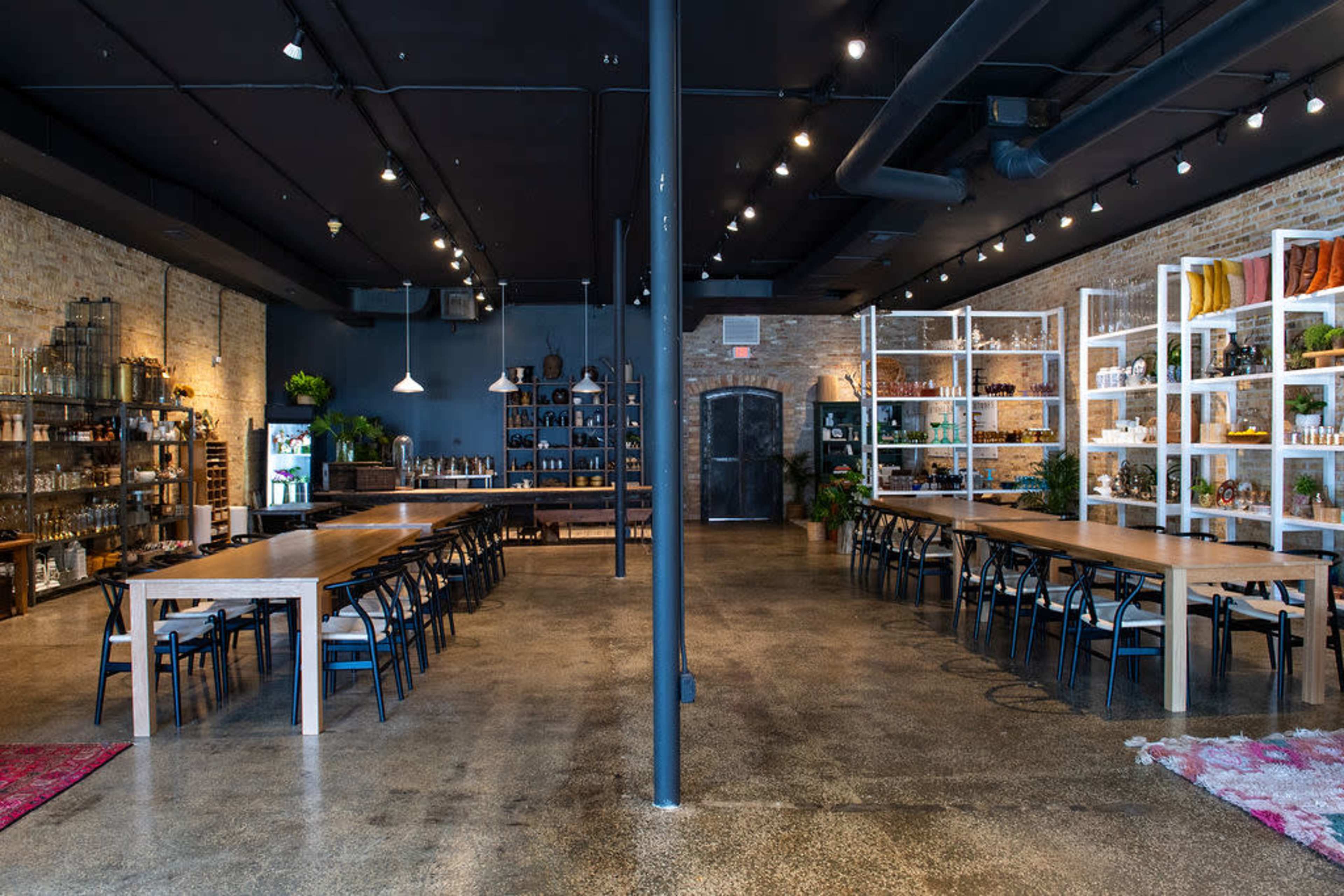 A spacious, modern interior of a restaurant or event space with long wooden tables, black chairs, and shelves displaying various bottles and decor against exposed brick walls.