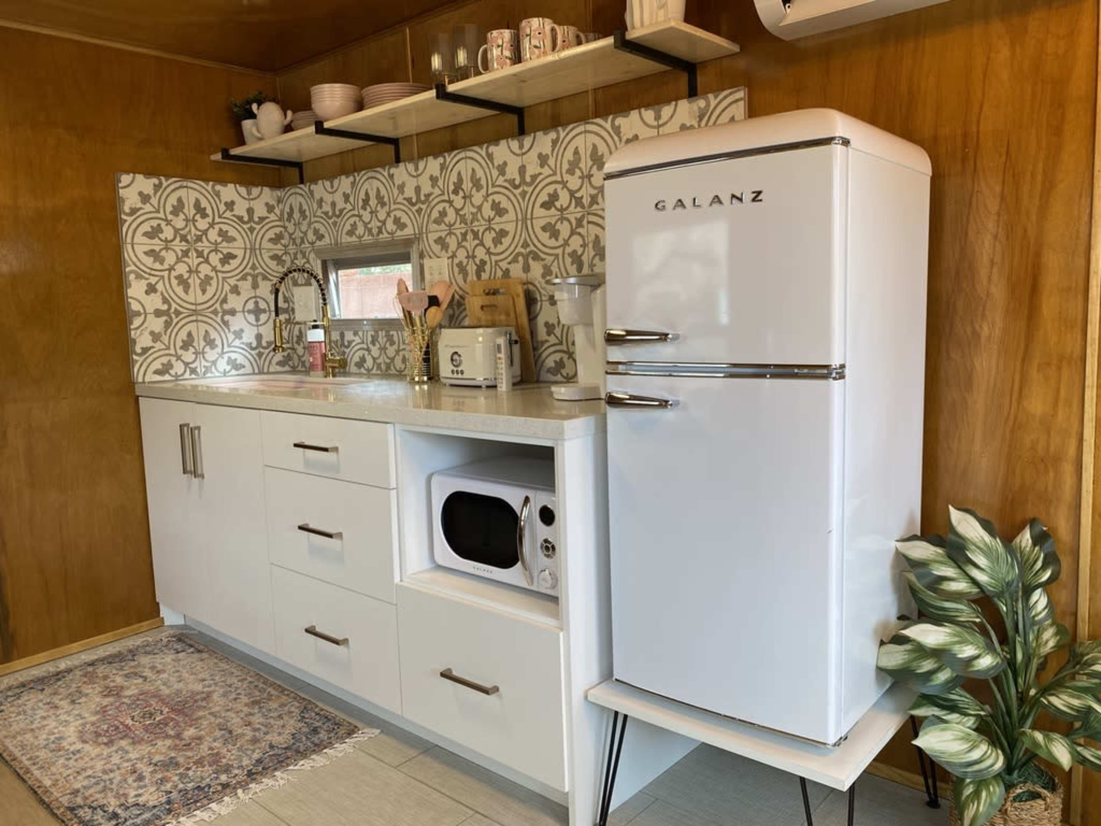 The image shows a small kitchen featuring a white retro-style refrigerator, a microwave under the counter, and patterned tile backsplash above a sink and countertop.