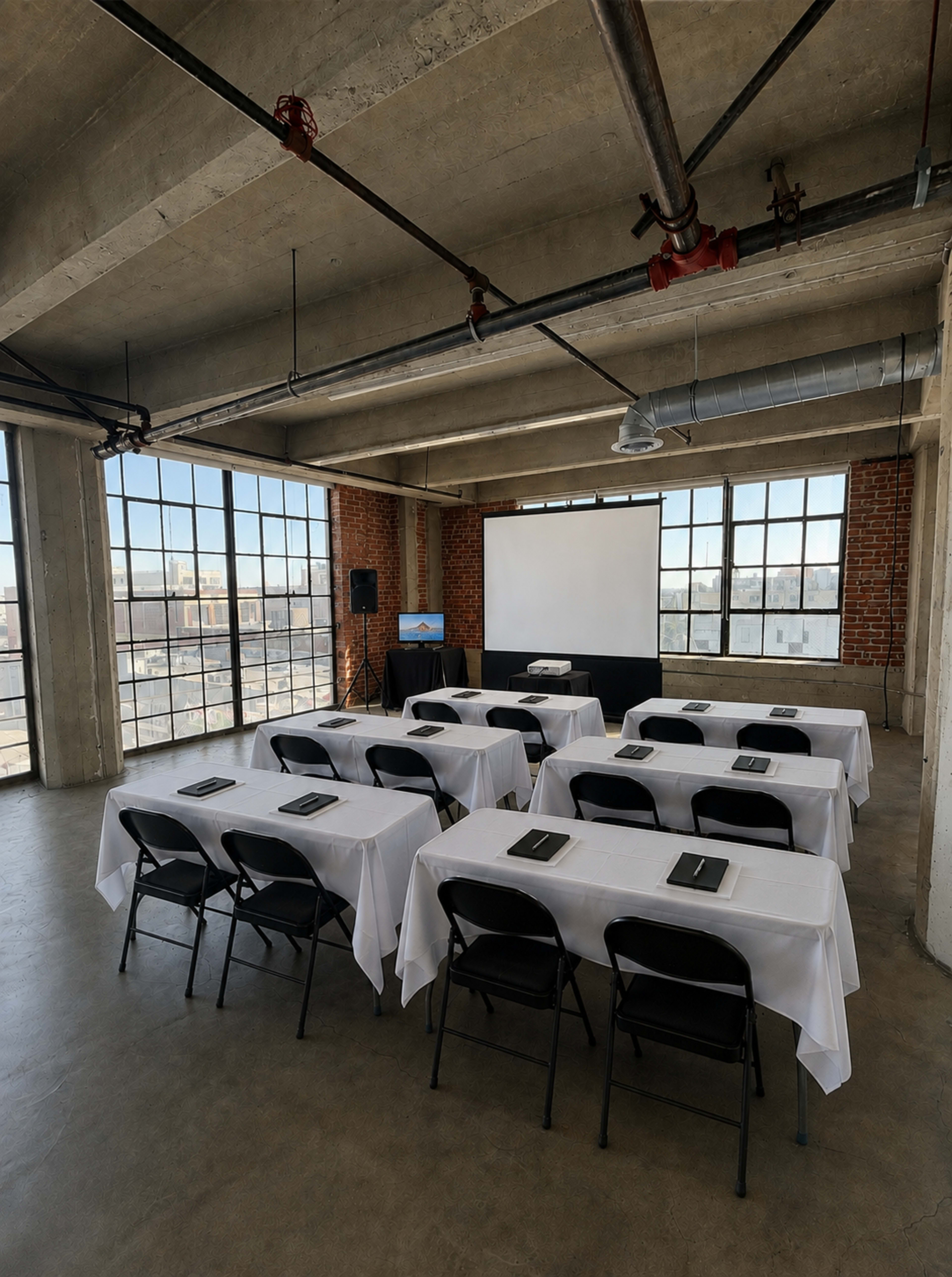 The image shows a spacious conference room set up with several rows of tables covered in white tablecloths and black chairs, featuring a projector screen and windows overlooking a cityscape.