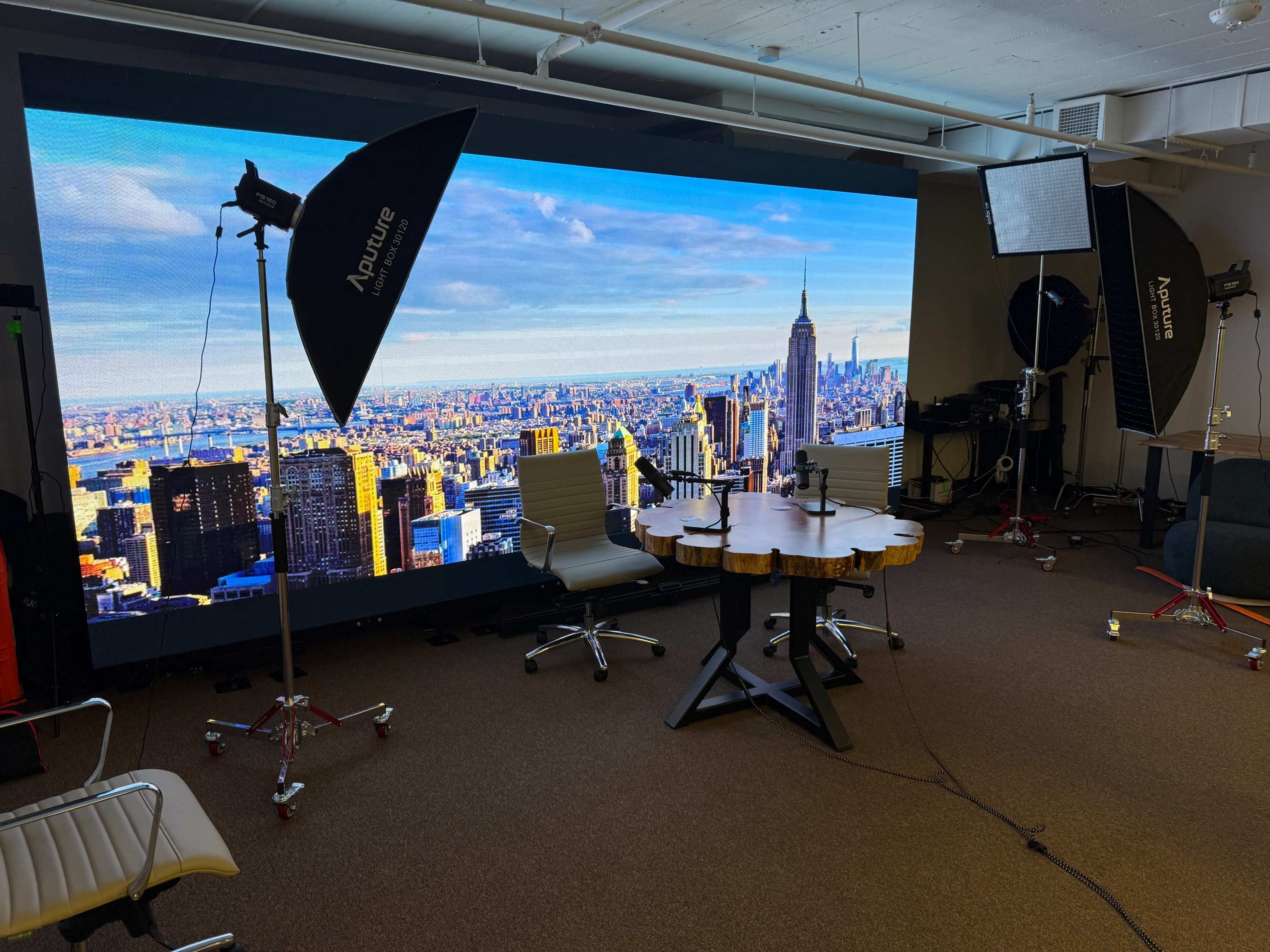 The image shows a studio setup with a large backdrop of a city skyline, two chairs, and lighting equipment arranged around a wooden table.