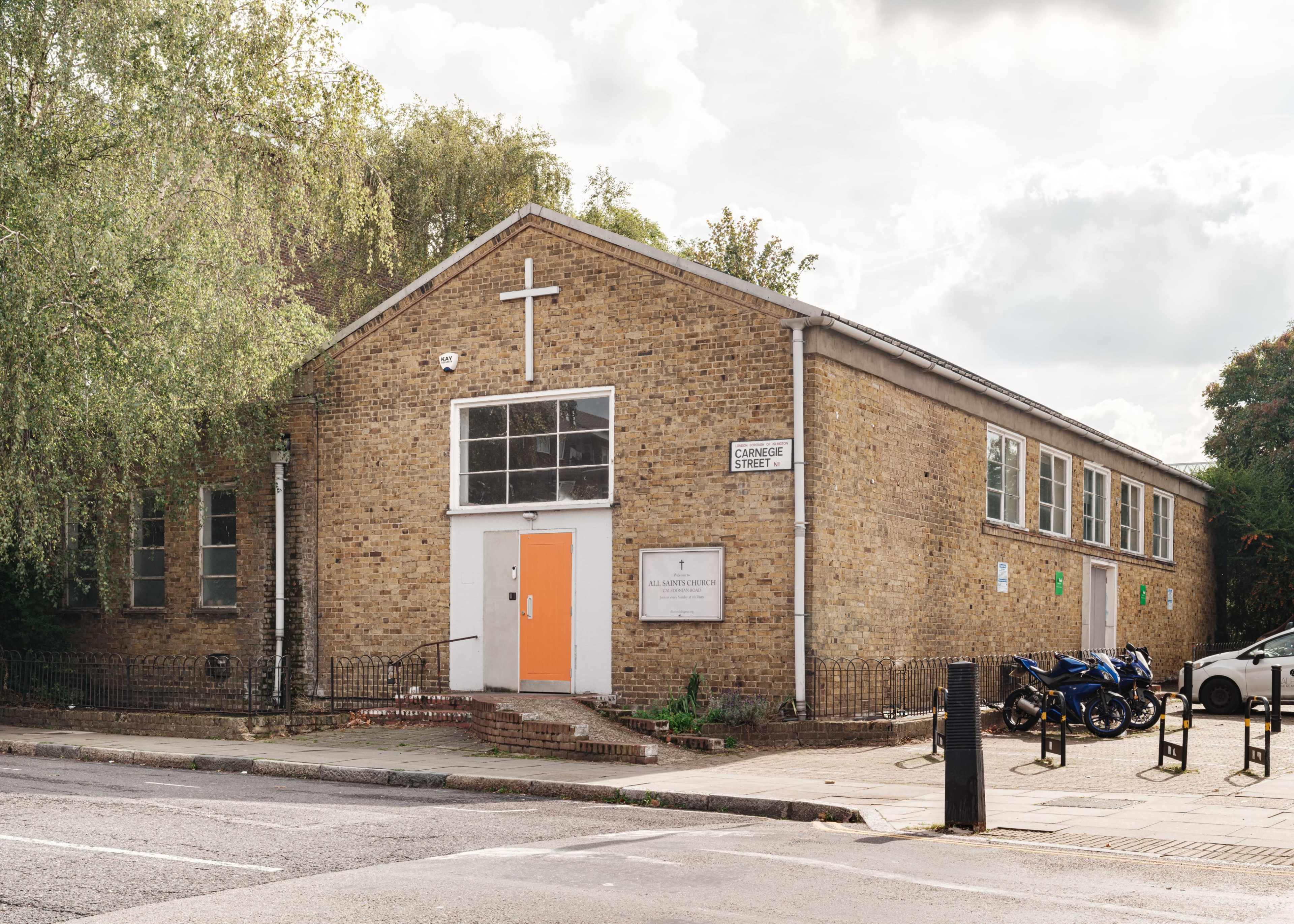 The image shows a brick building with a cross on the facade, an orange door, and multiple windows, located at the corner of a street with parked motorcycles nearby.