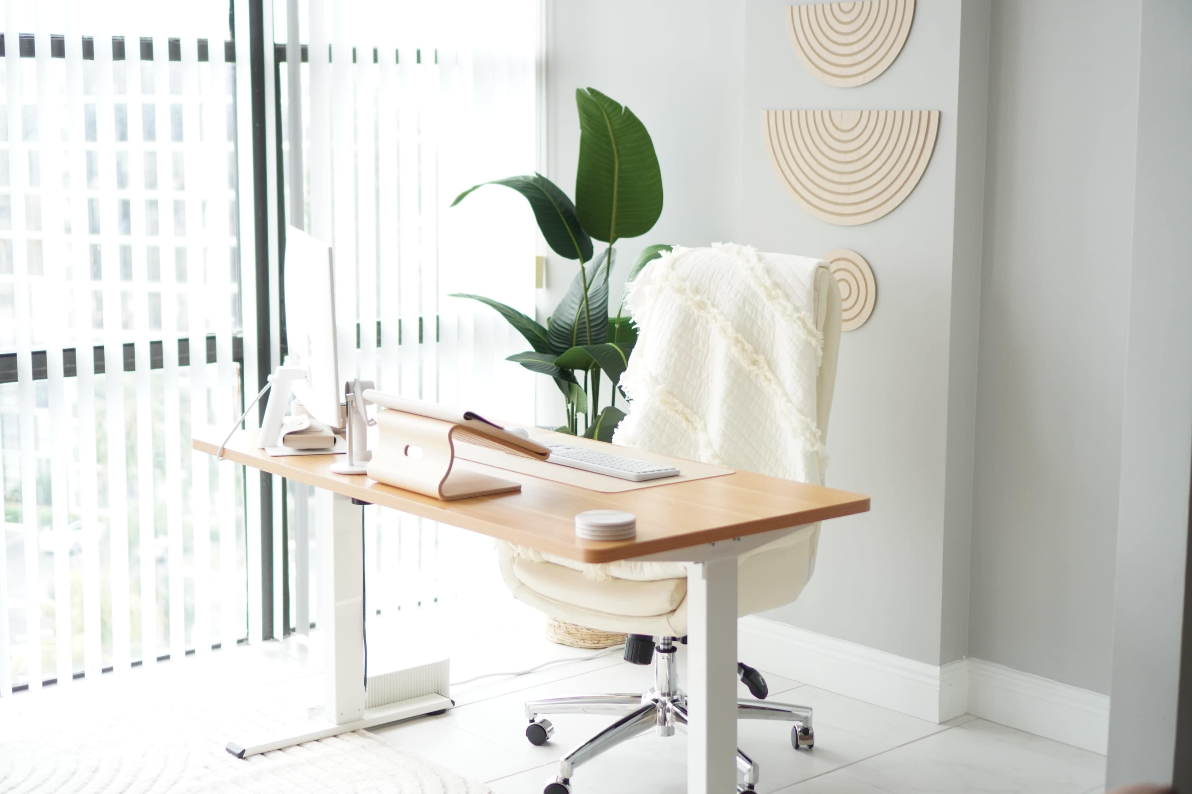 A modern home office features a wooden desk with a computer, a white chair, and a potted plant beside a window with vertical blinds.